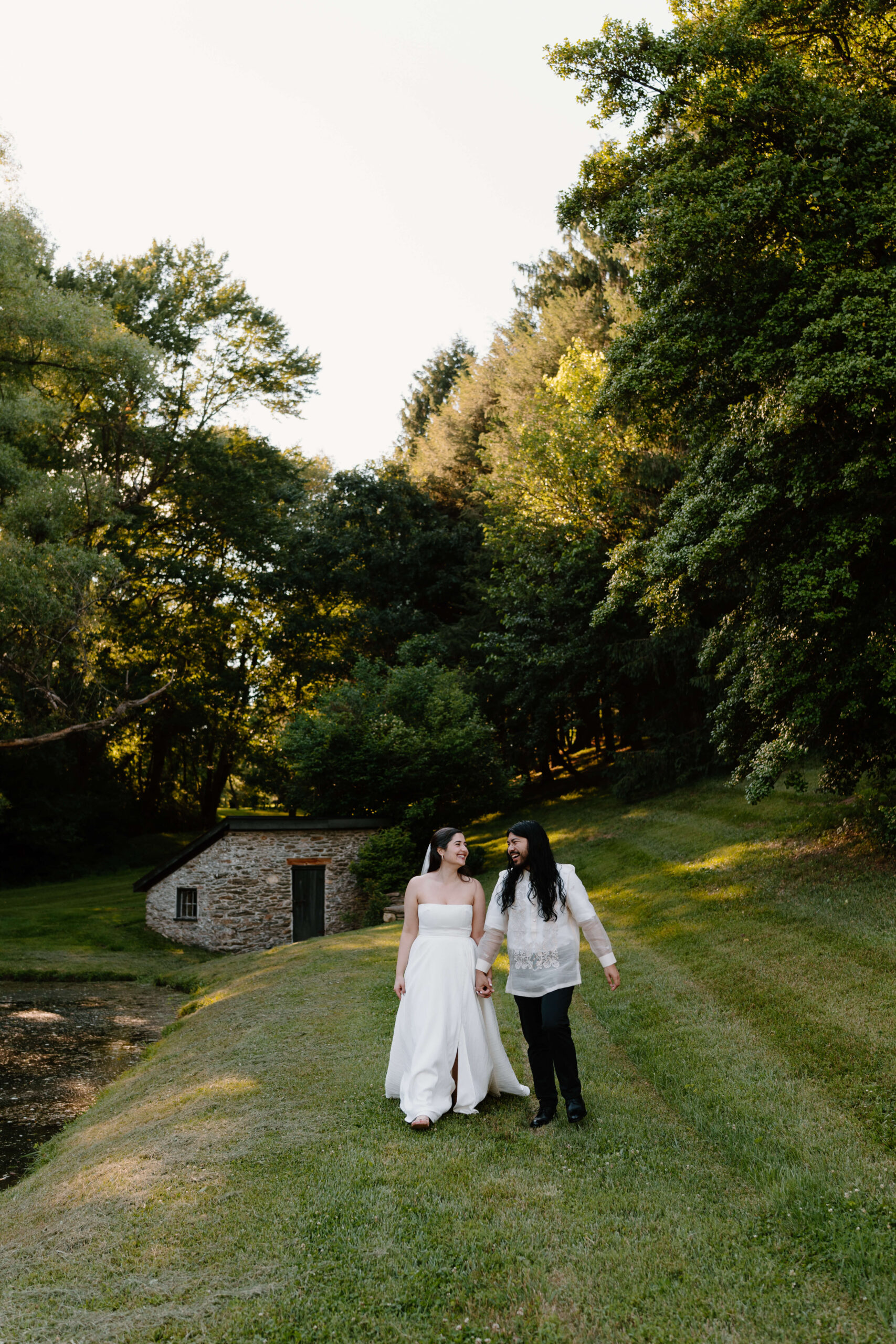 a bride and groom (both long brown hair) walking toward the camera in a green grassy field surrounded by tall trees. She is wearing a strapless white gown, and he is wearing a white shirt and black pants.