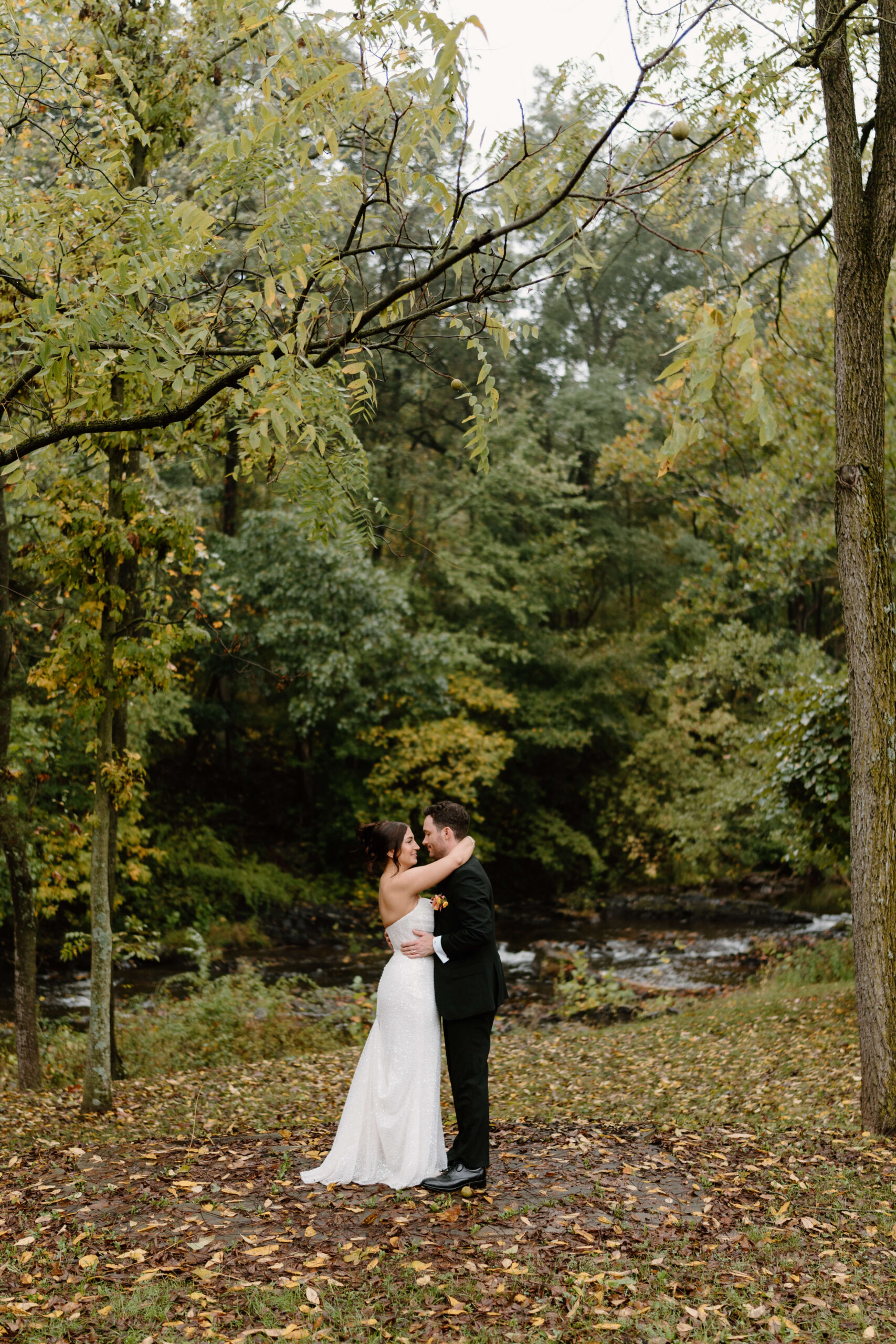 bride (brown hair in an updo) and groom (brown hair, black suit and a boutonniere) embracing in front of a river, surrounded by fall leaves on a rainy day