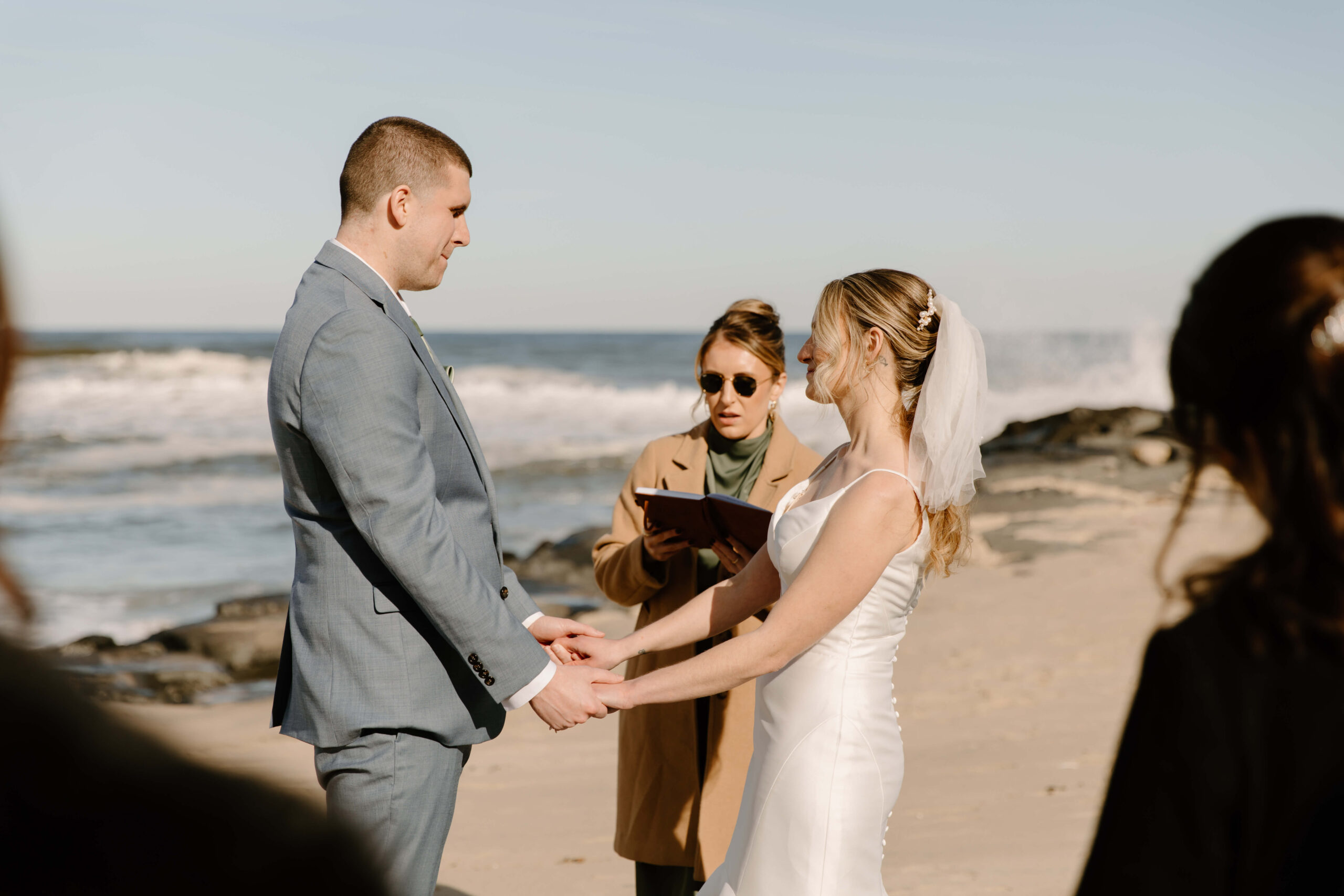 bride and groom holding hands during their elopement ceremony at the beach, while their officiant performs the ceremony wearing a jacket. The ocean waves crash behind them on the rocks