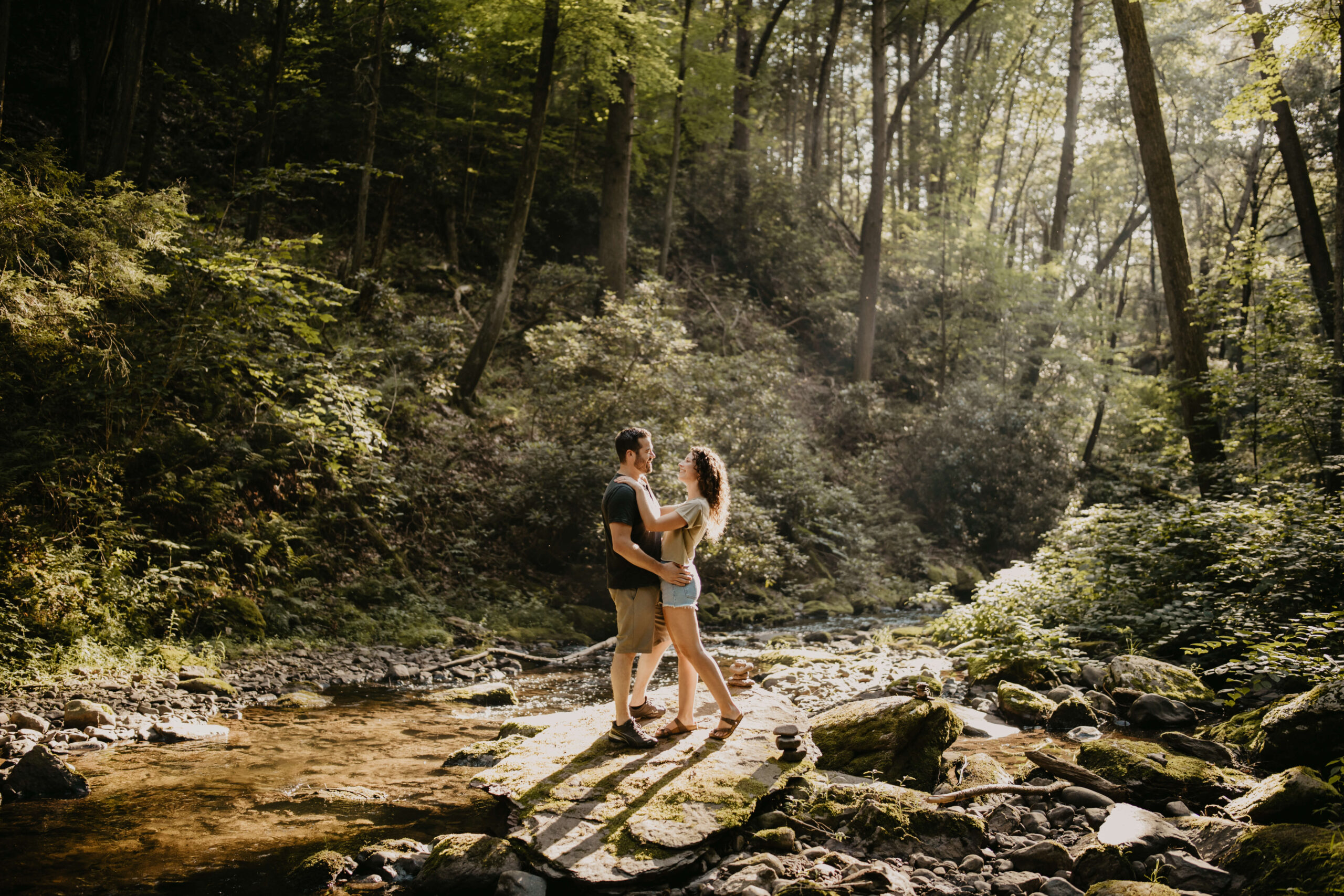 a couple embracing and smiling at each other while on a hike, standing on a large rock in the middle of a creek illuminated in sunlight and surrounded by bright green summer foliage