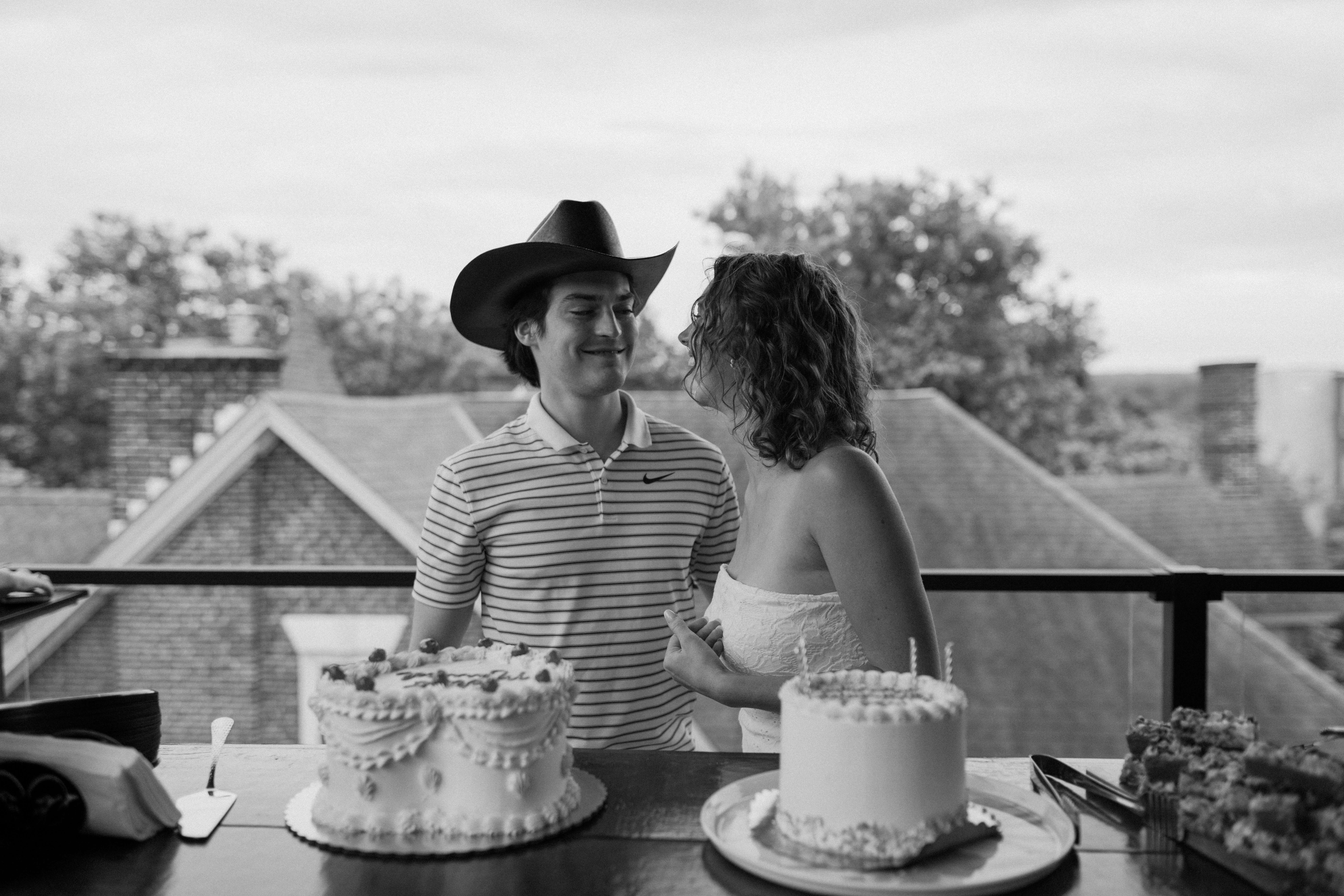 black and white image of a bride (strapless white dress) and groom (striped polo shirt, cowboy hat) smiling at each other while standing over their vintage decorated wedding cake