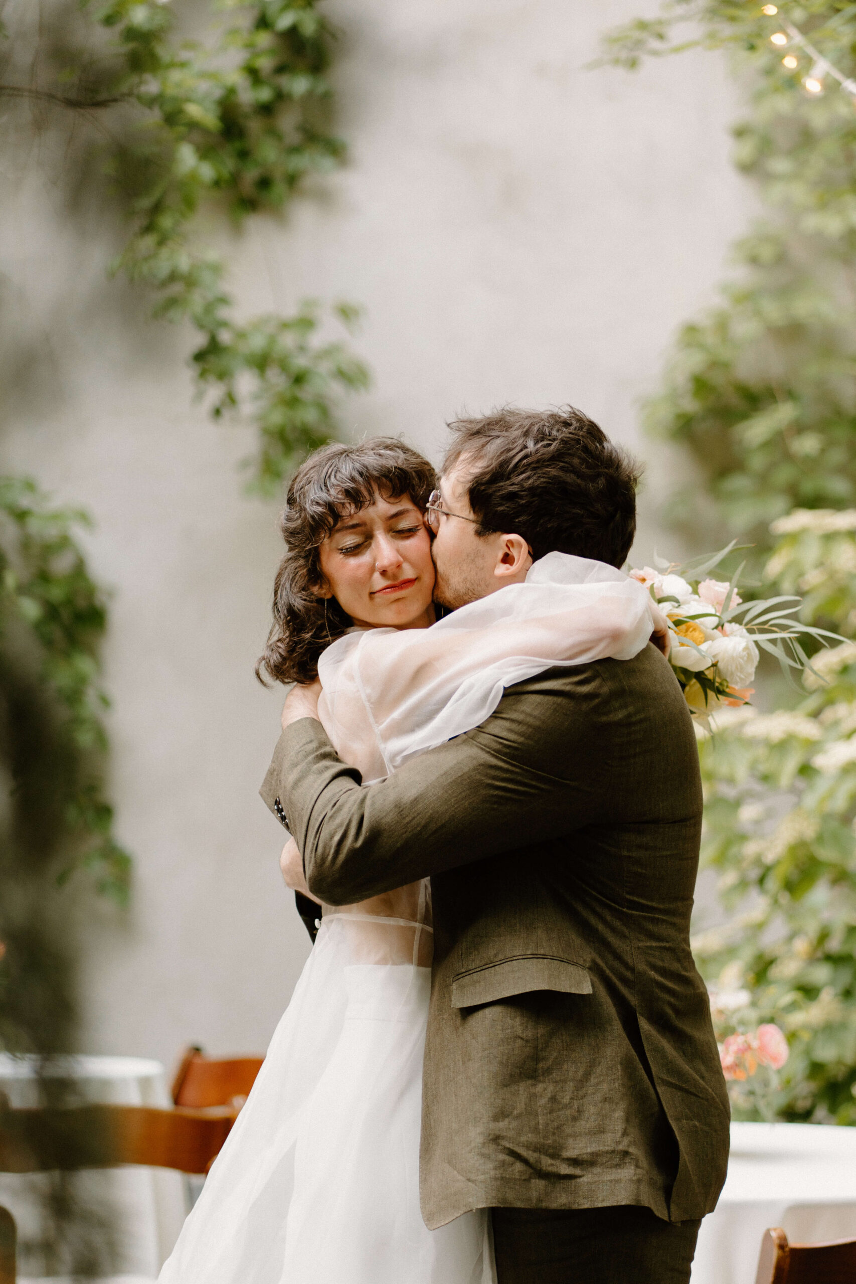 a groom (brown hair, olive green suit jacket) hugging and kissing his bride (sheer white dress) on the cheek as she closes her eyes and holds back tears