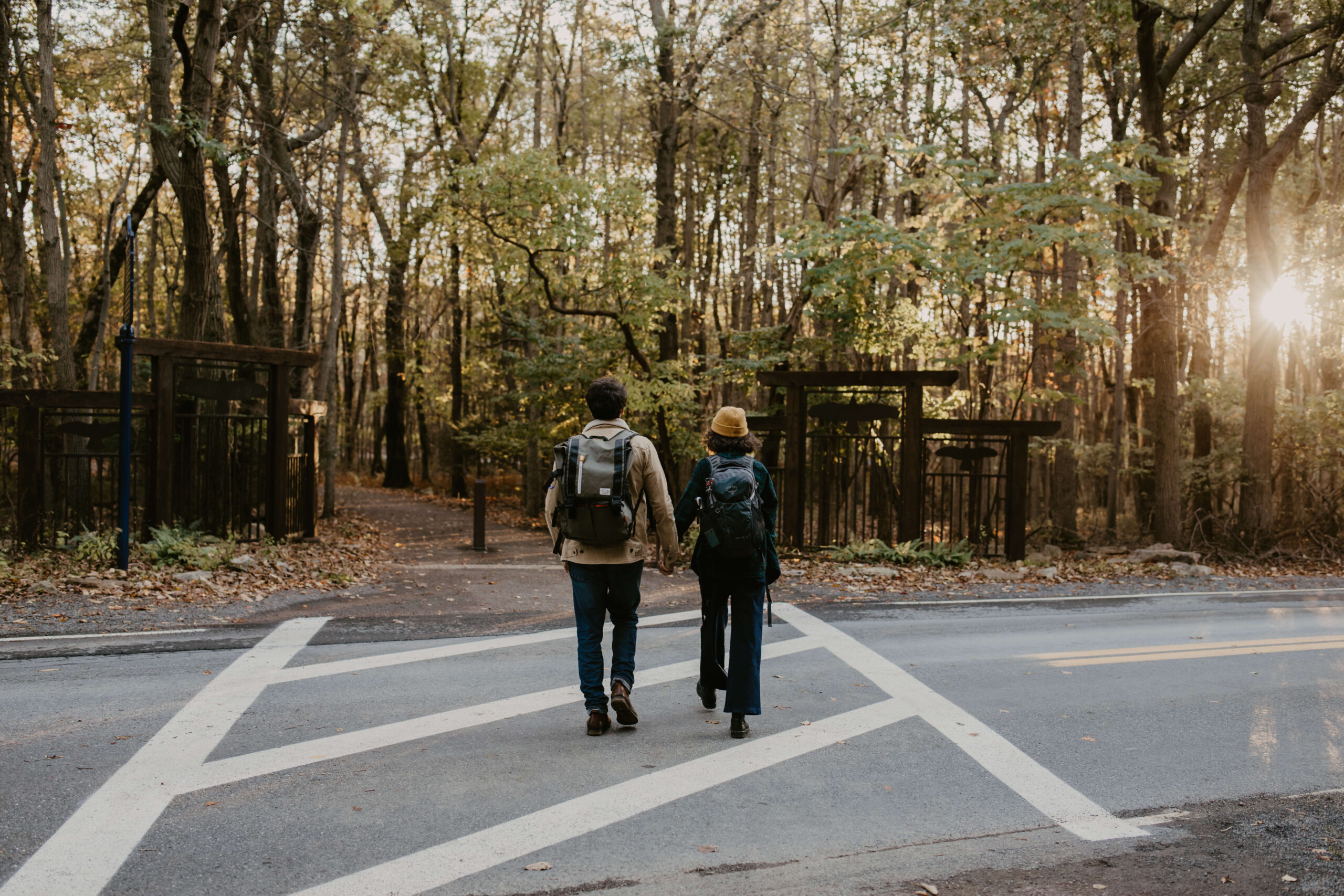 a couple holding hands and walking across the road, away from the camera, back to a parking lot after a hike, wearing boots and backpacks