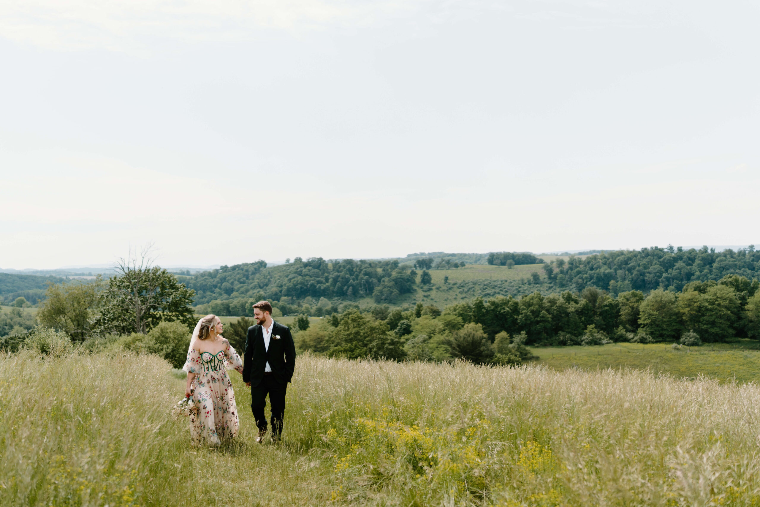 bride and groom holding hands and walking through a field of tall green grass while smiling at each other, rolling fields extending in the distance