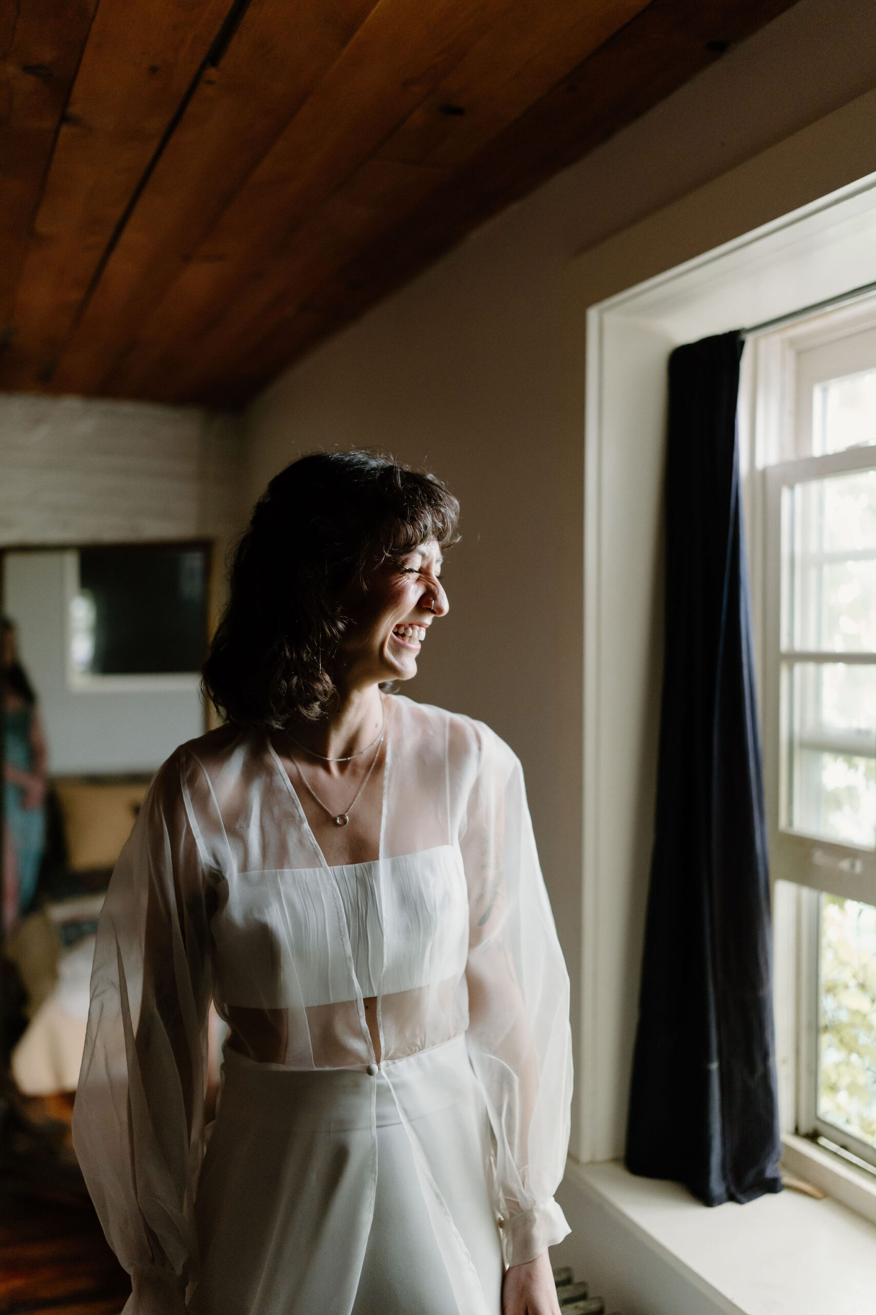 bride in a two piece wedding gown and a sheer cape with a curly short haircut, laughing toward a window