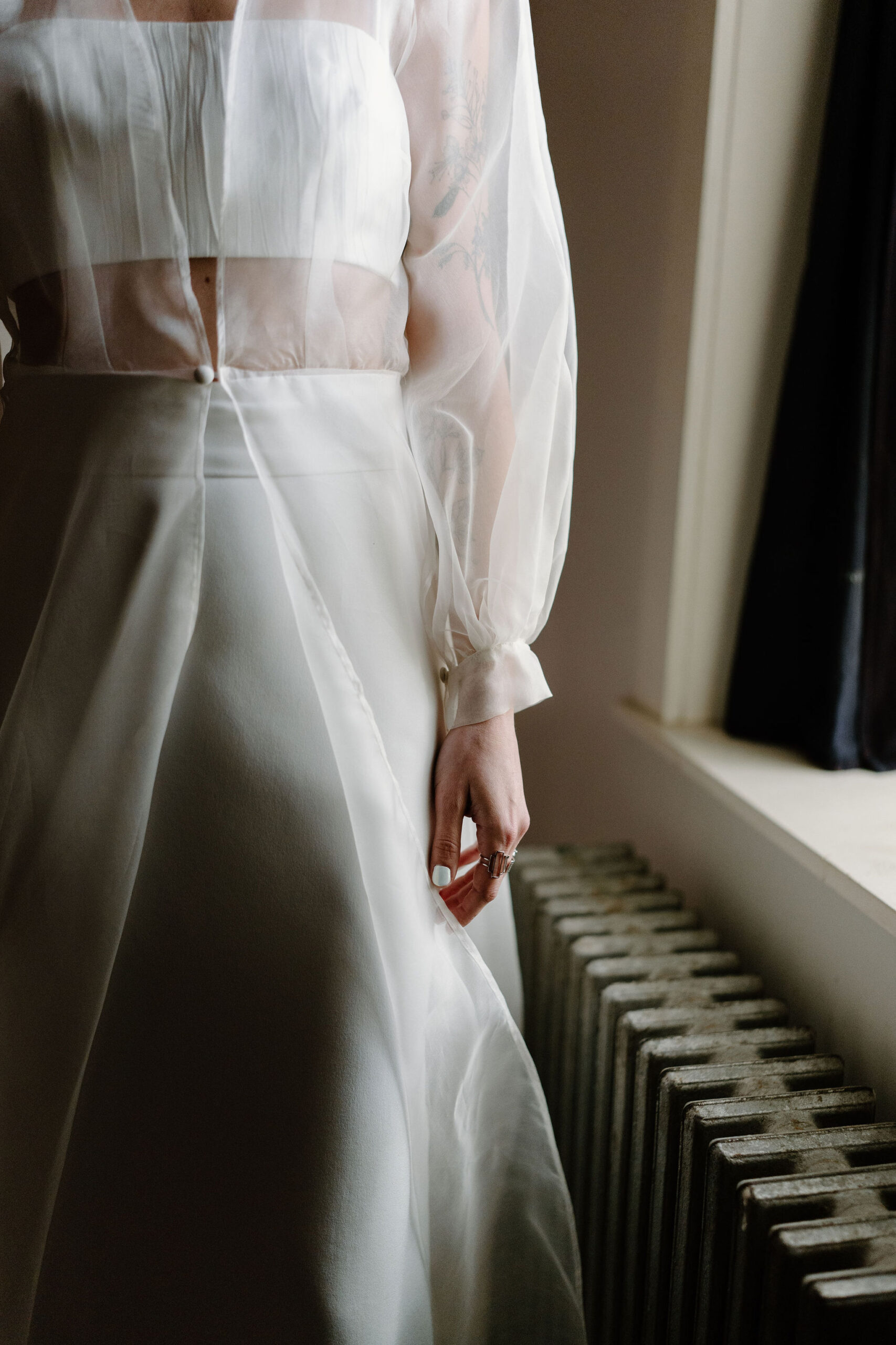 close up image of a bride wearing a two piece cropped spaghetti strap top, a simple skirt, and a sheer cape with one button at her waist, her hand held at her side with lots of rings