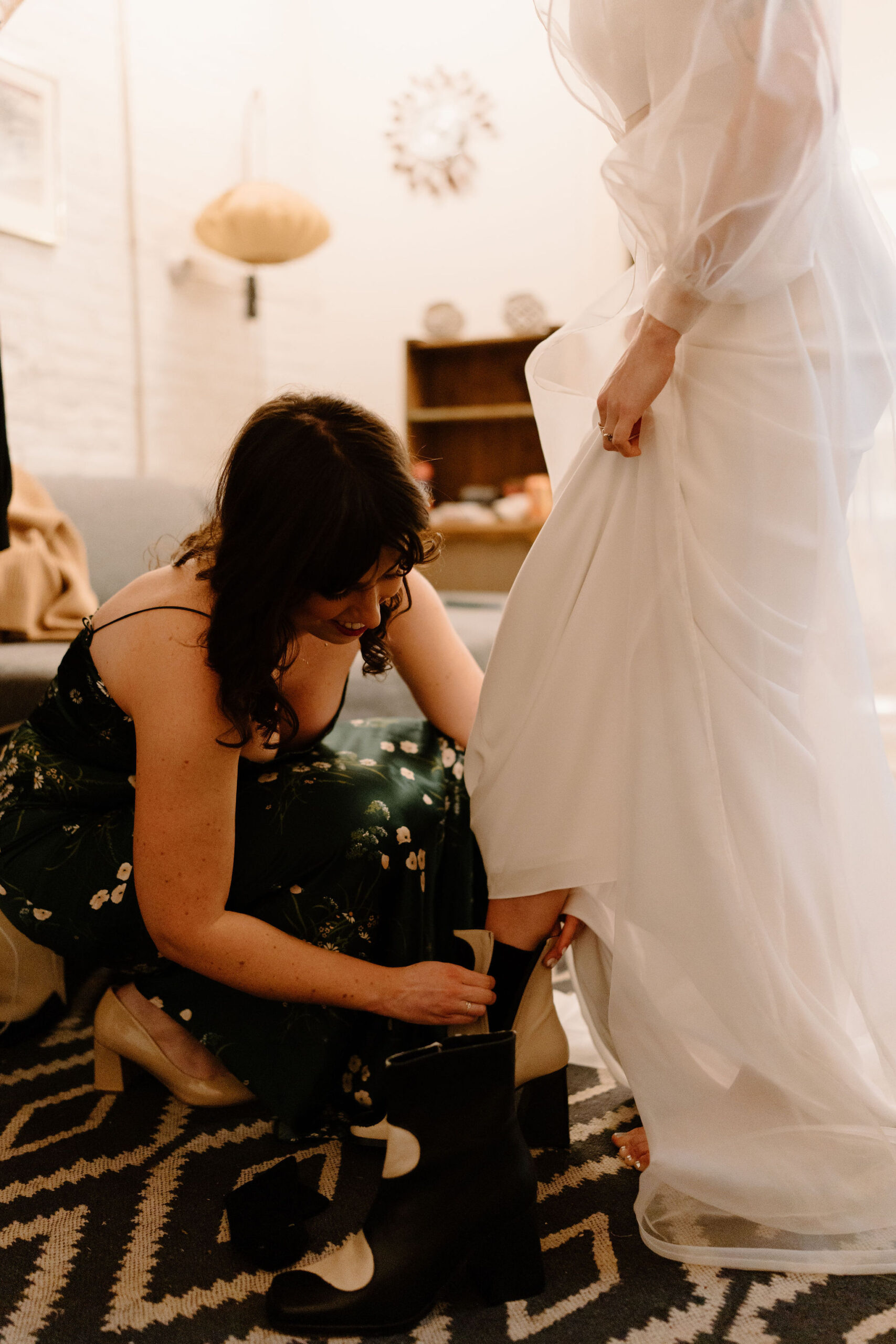 a bride stepping into her wedding boots, holding up her skirt while her friend helps her zip them up