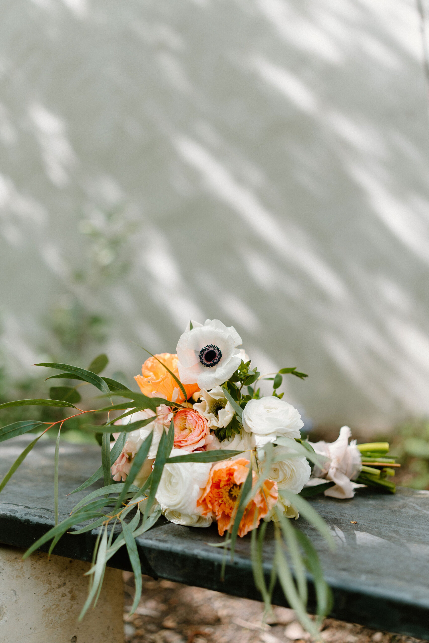 bridal bouquet of greenery, poppies, eucalyptus, and other orange flowers, positioned on a bench outdoors against a white wall with dappled shadows