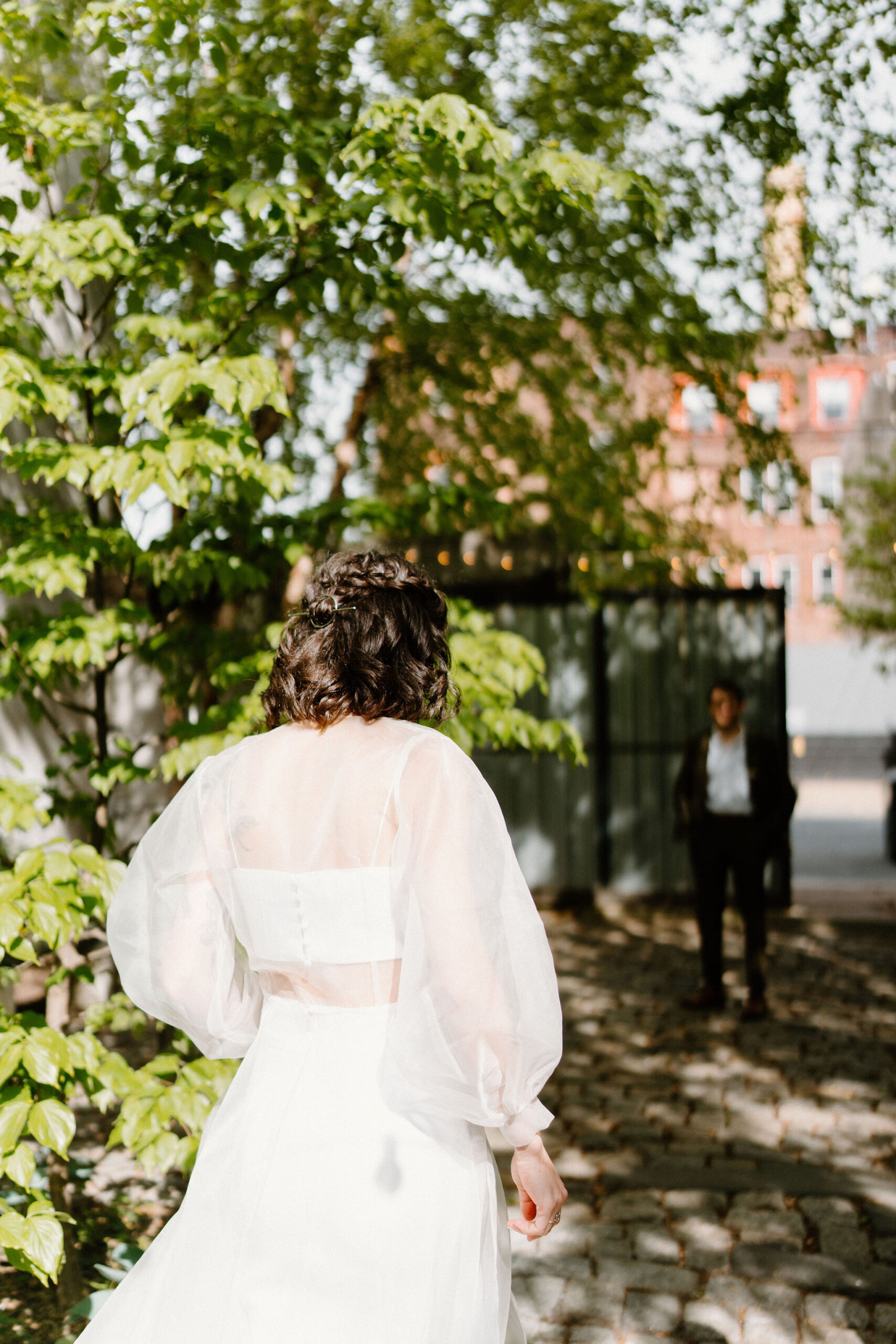 bride (brown hair, sheer cape, two piece gown) walking up to her groom (brown hair, green suit) in a lush green cobblestone courtyard