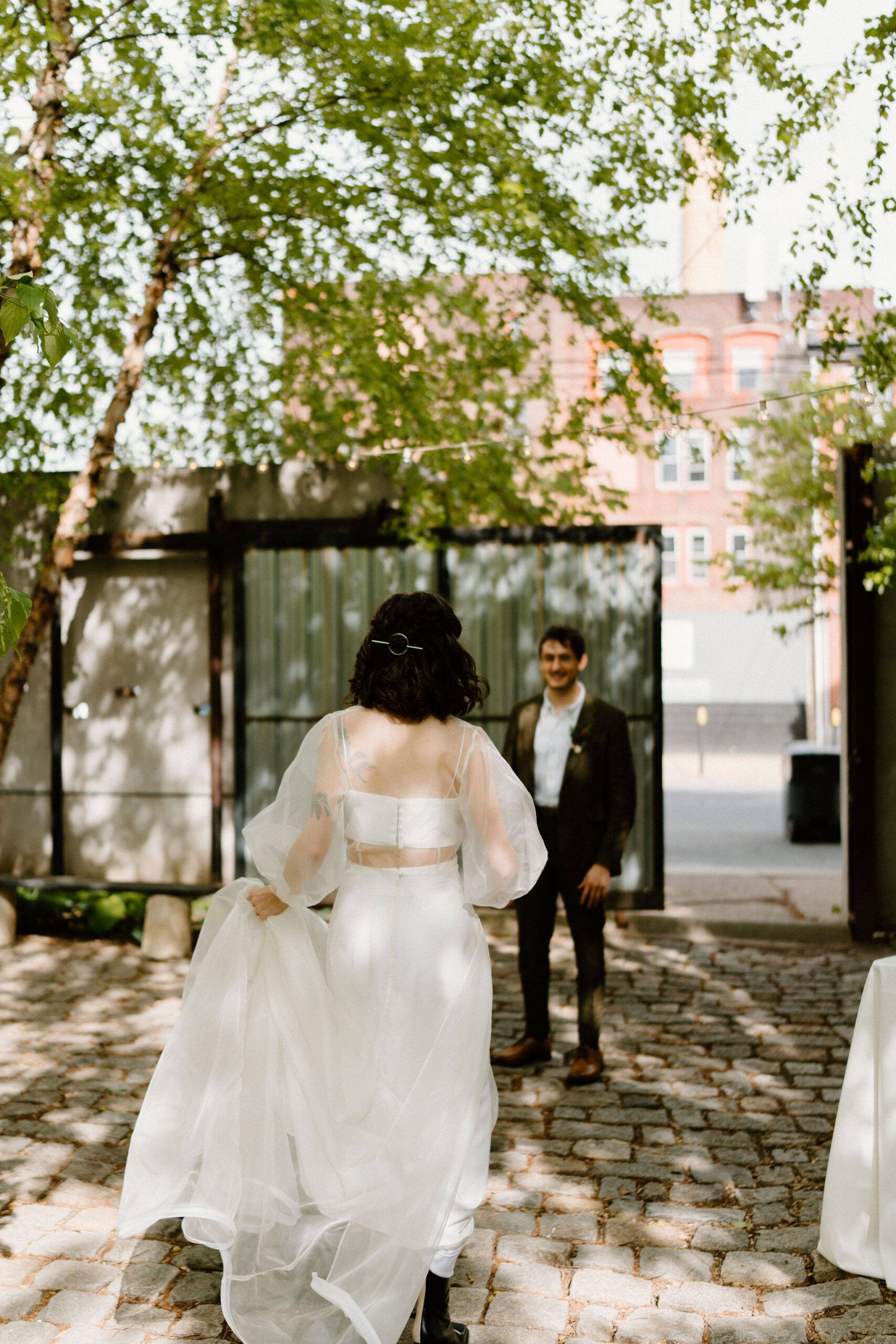 bride (brown hair, sheer cape, two piece gown) walking up to her groom (brown hair, green suit) in a lush green cobblestone courtyard