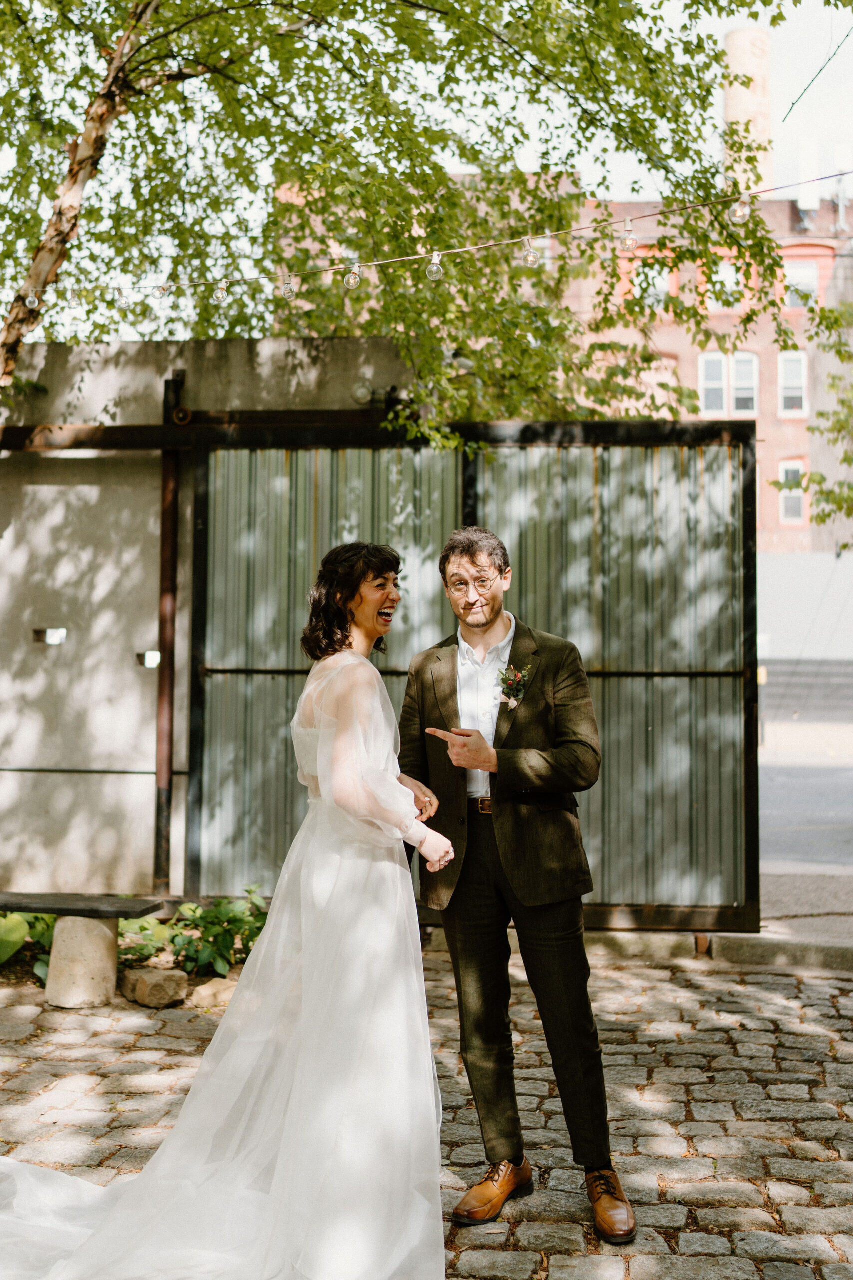 bride (brown hair, sheer cape, two piece gown) and groom (brown hair, green suit) in a lush green cobblestone courtyard, her laughing at him and him smirking at the camera, pointing at her