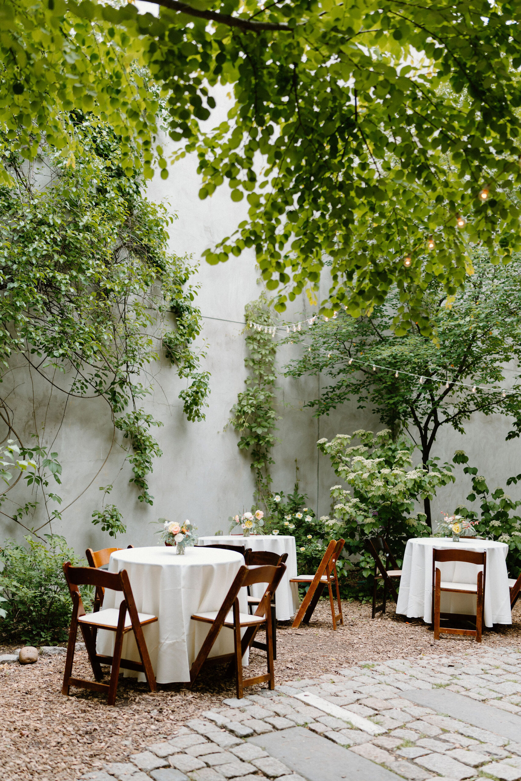 wide image of three tables set up with wooden chairs, white tablecloths, and a small bouquet of flowers, inside a lush green courtyard garden with cobblestone