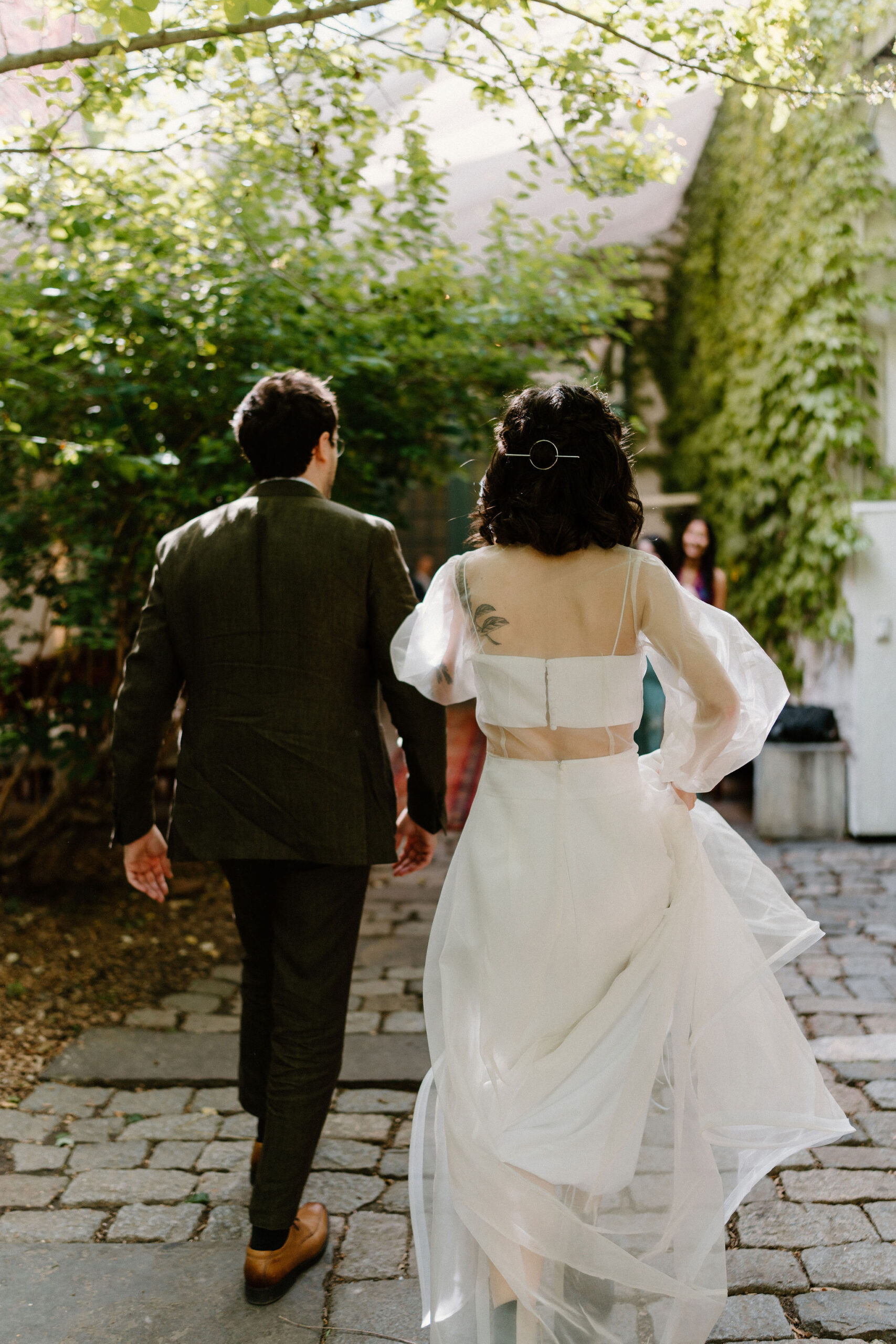 bride and groom walking through a lush green cobblestone courtyard toward their friends, away from camera