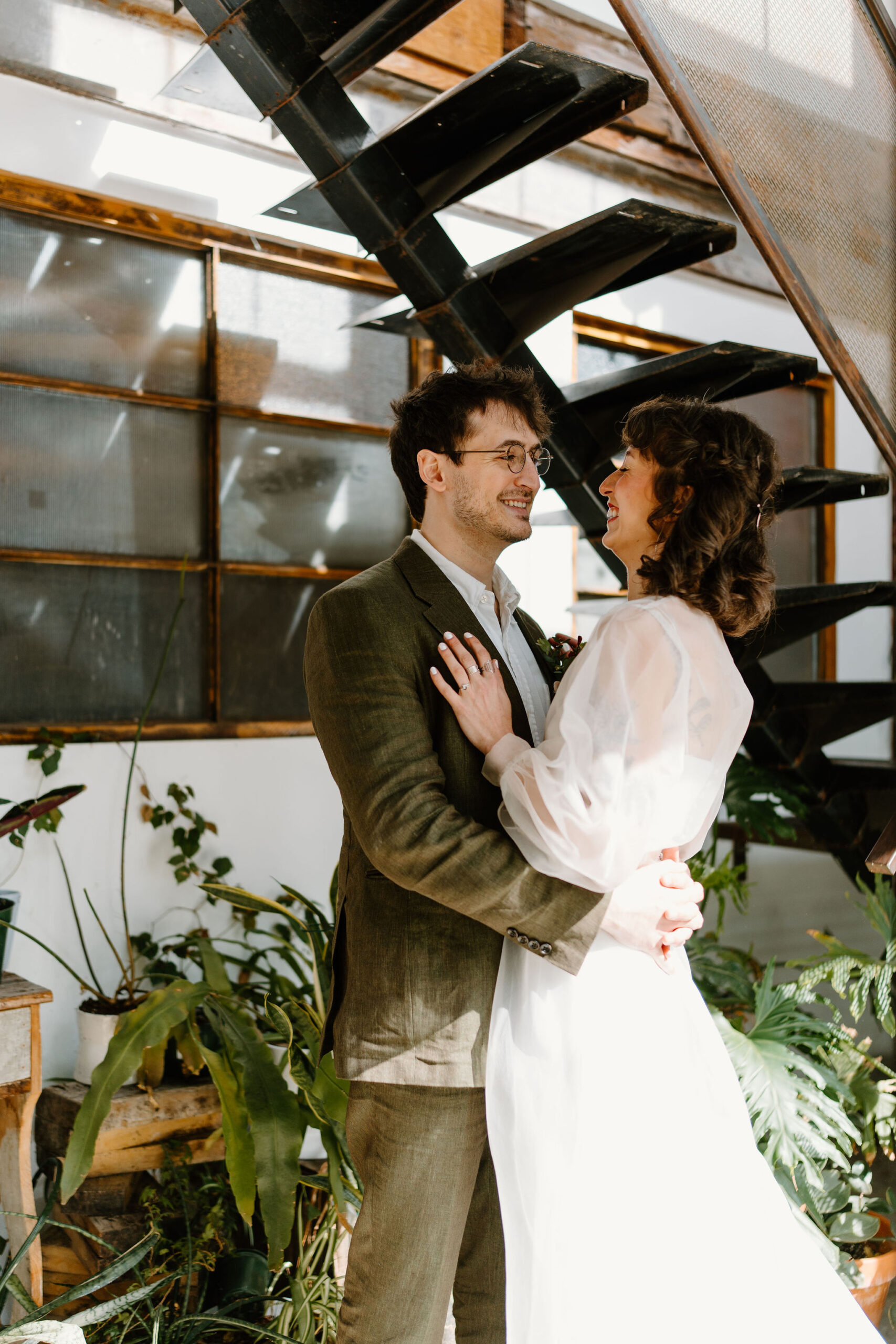 close up image of a bride (white, sheer dress) and groom (linen olive suit) facing each other, embracing and laughing, in front of a wall full of plants and an industrial staircase