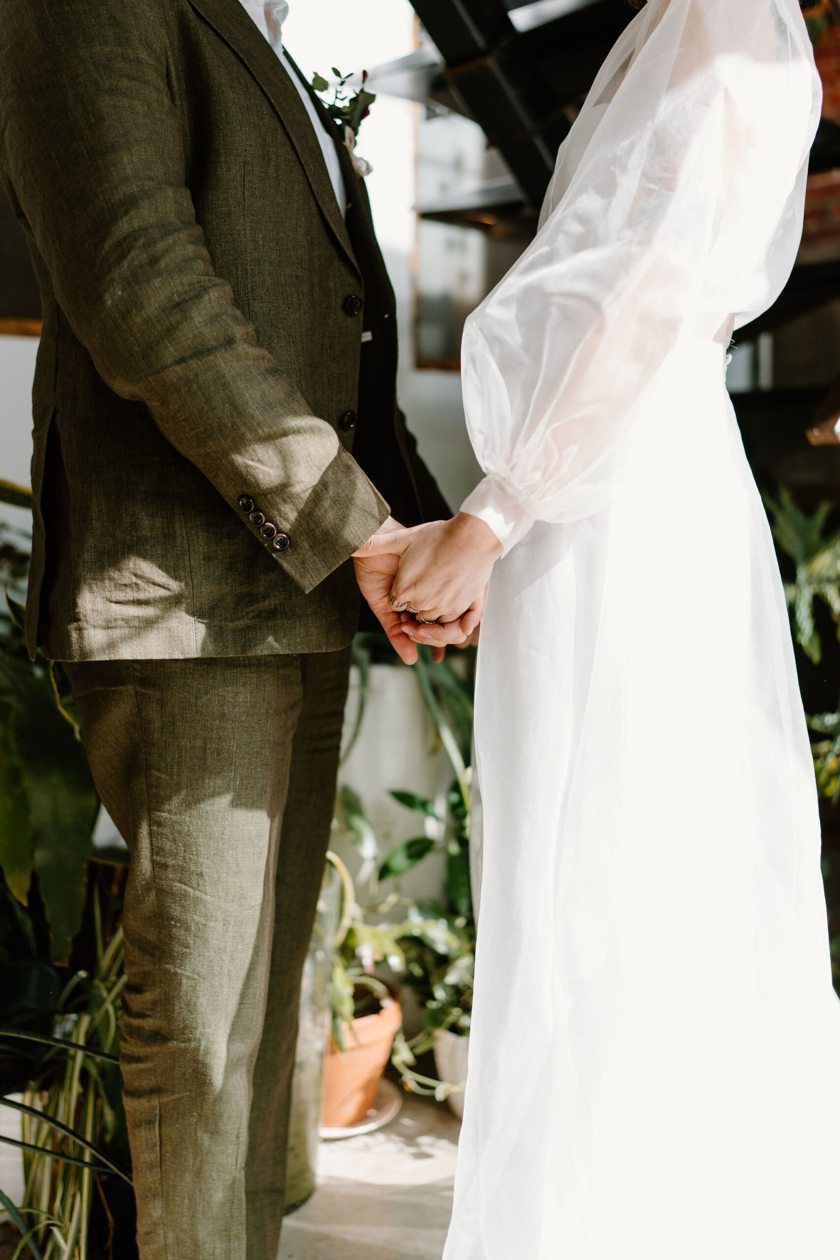 close up image of a bride (white, sheer dress) and groom (linen olive suit) facing each other and holding hands  in front of a wall full of plants and an industrial staircase