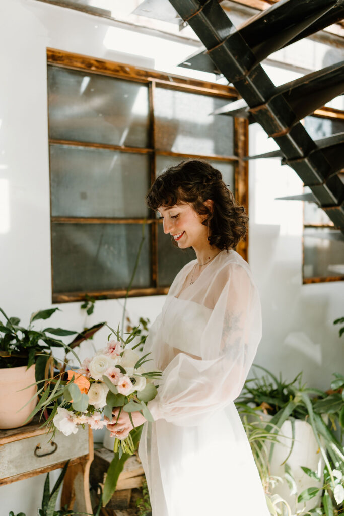 bride (short brown curly hair, bangs) holding her bouquet and smiling softly down at her flowers in front of a wall full of plants and an industrial staircase