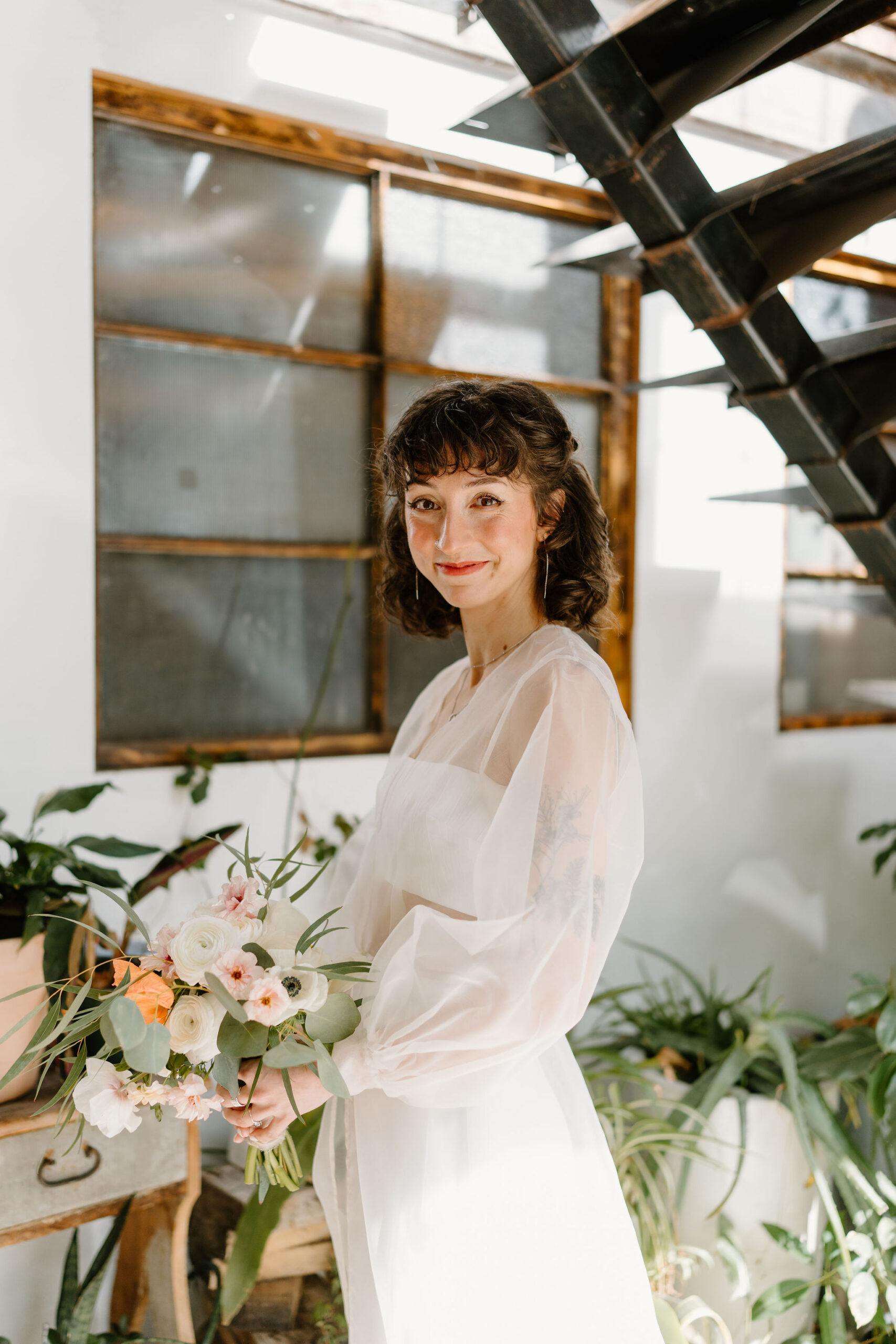 bride (short brown curly hair, bangs) holding her bouquet and smiling softly toward the camera in front of a wall full of plants and an industrial staircase