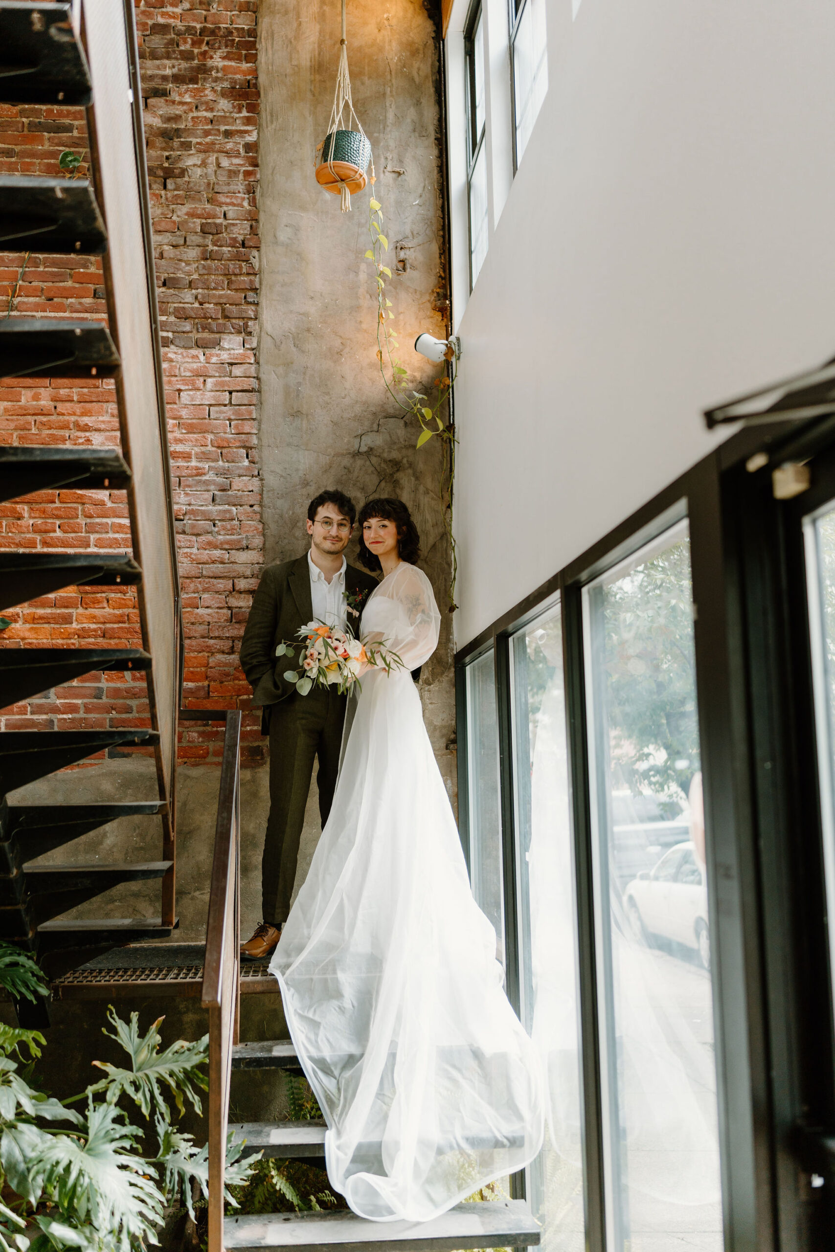 bride and groom standing on an industrial steel staircase in front of a brick wall, smiling at the camera. the bride is holding a bouquet while her train falls down the stairs