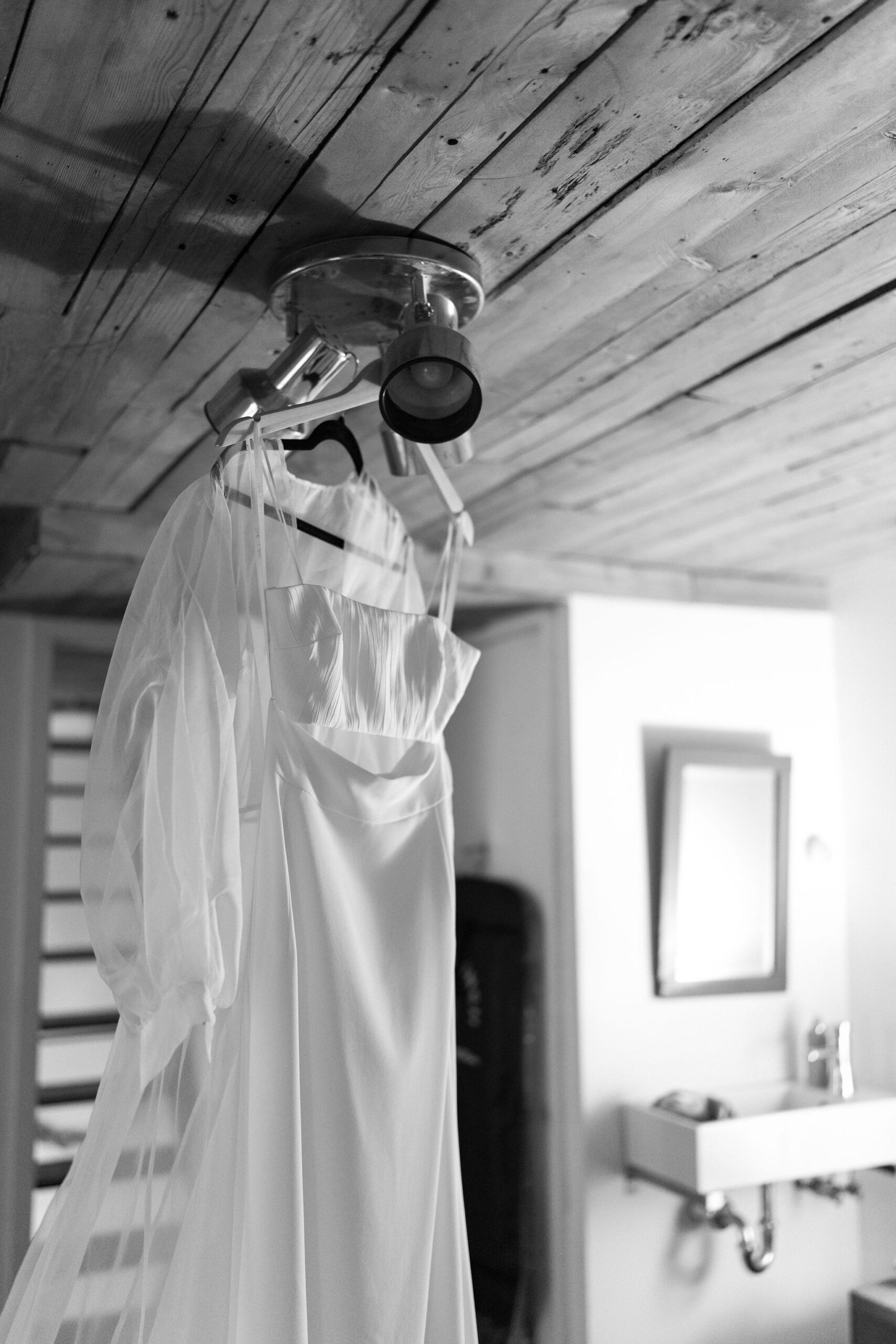 black and white image of a two-piece wedding dress and a cape, hanging from a ceiling light fixture in a bedroom with a sink in the background