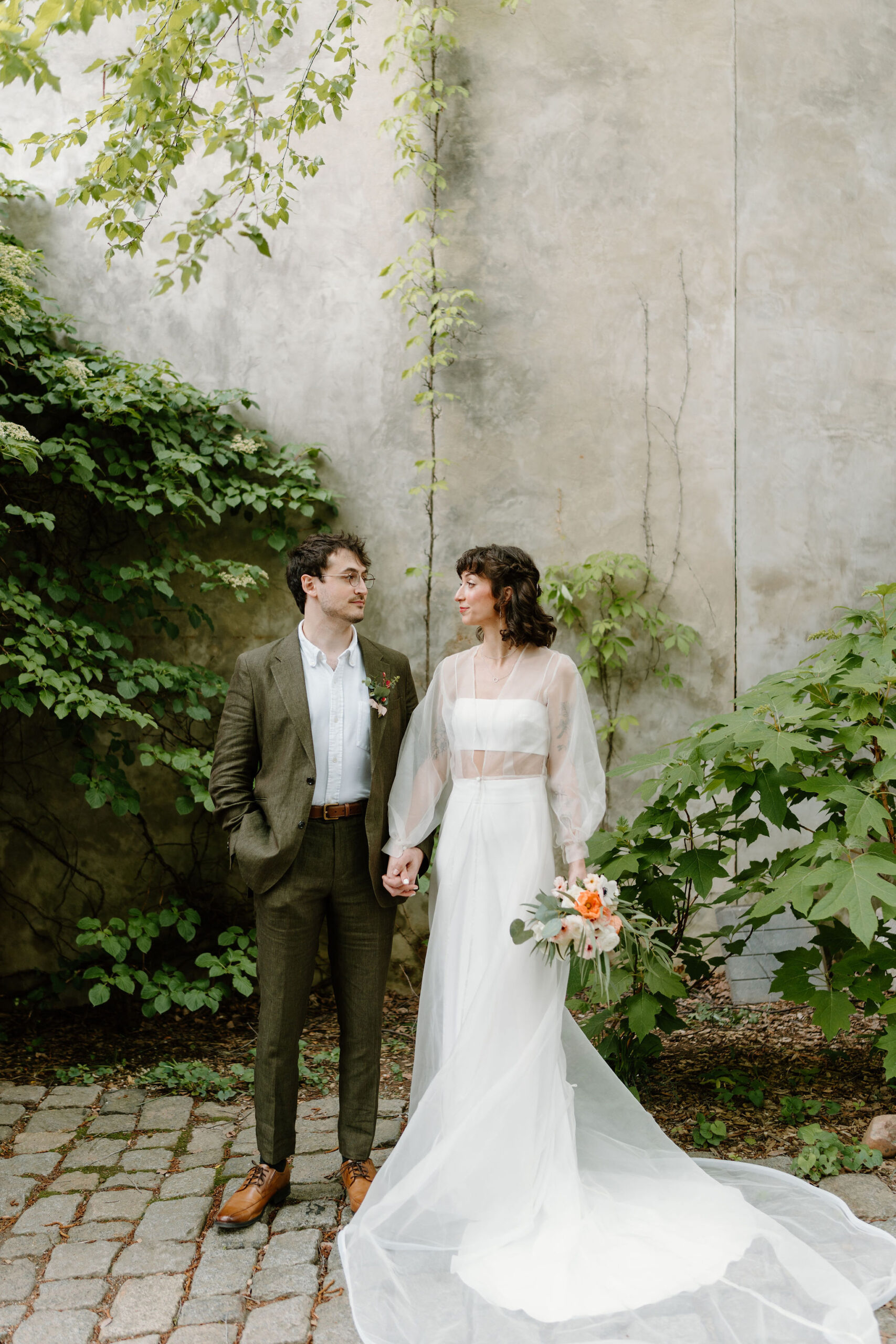 bride (two piece cropped dress with a sheer long sleeve cape, curly short brown hair with bangs) holding her bouquet at her side and holding groom's hand (brown hair, olive linen suit, brown shoes), smiling softly at each other in a lush green cobblestone courtyard