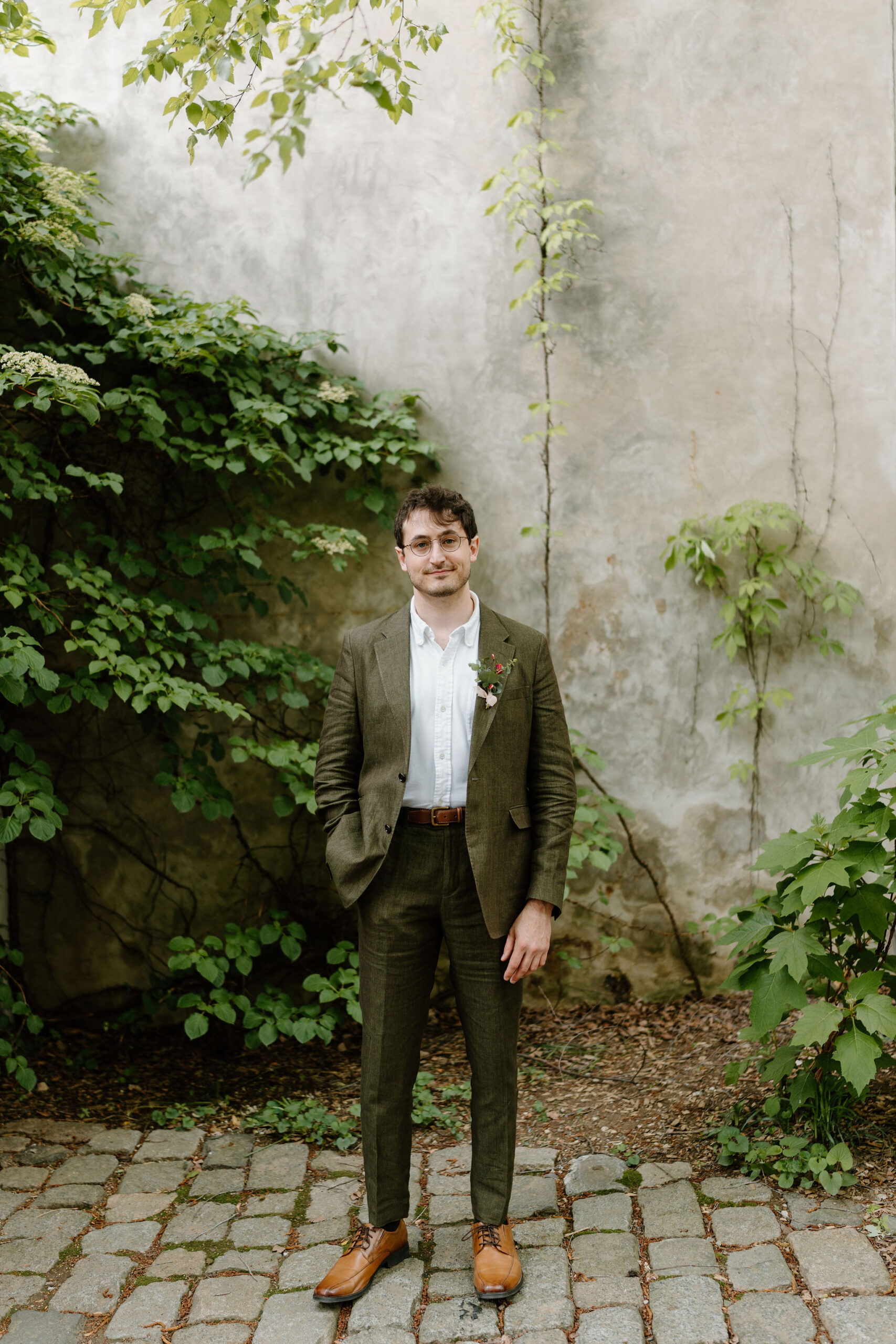 Groom (brown hair, glasses, white button down shirt, linen olive suit, and brown dress shoes) in a lush green cobblestone courtyard, smiling softly at the camera with his hand in his pocket