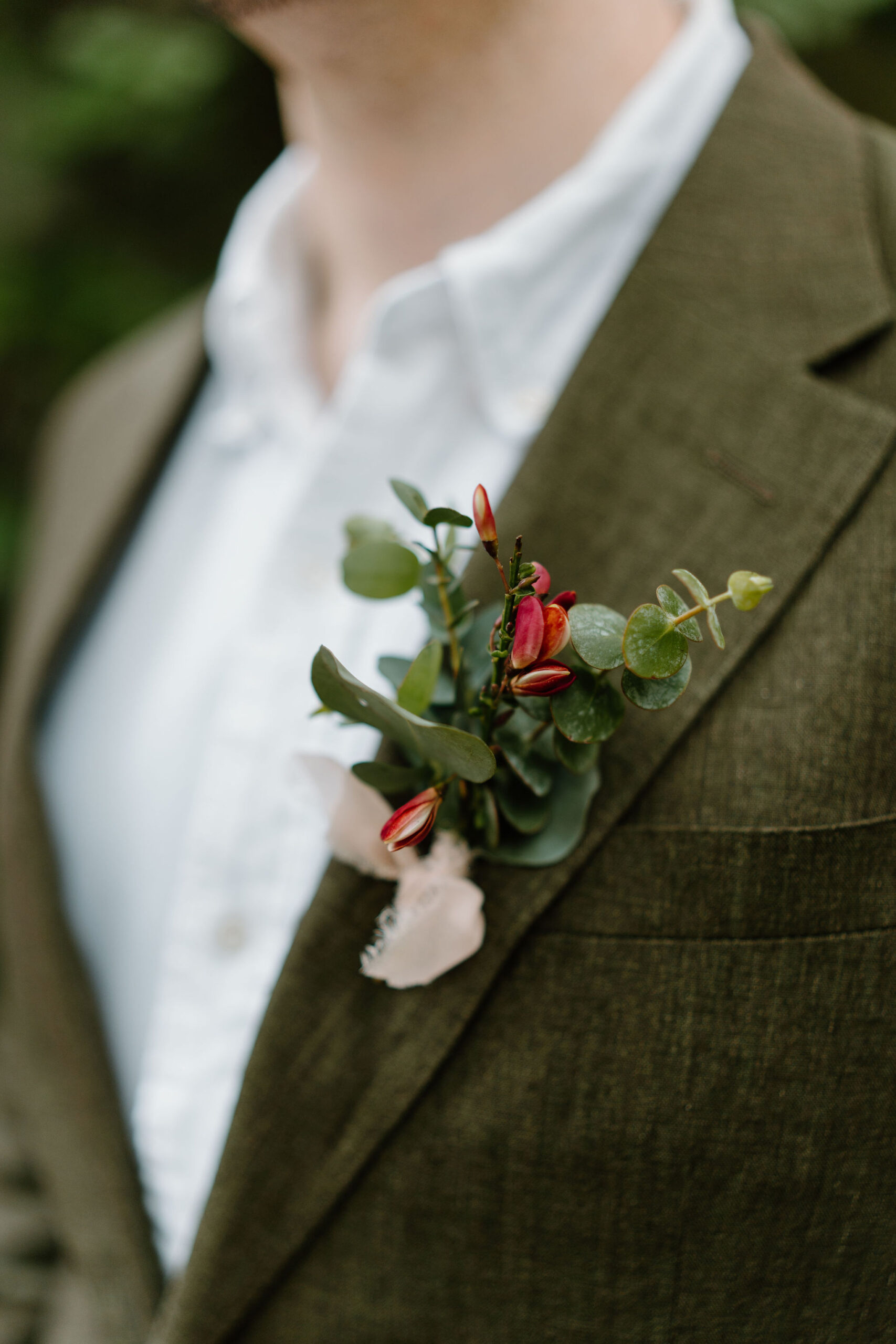 close up of a groom (white button down shirt, olive green linen suit) wearing a boutonniere with eucalyptus and small red buds