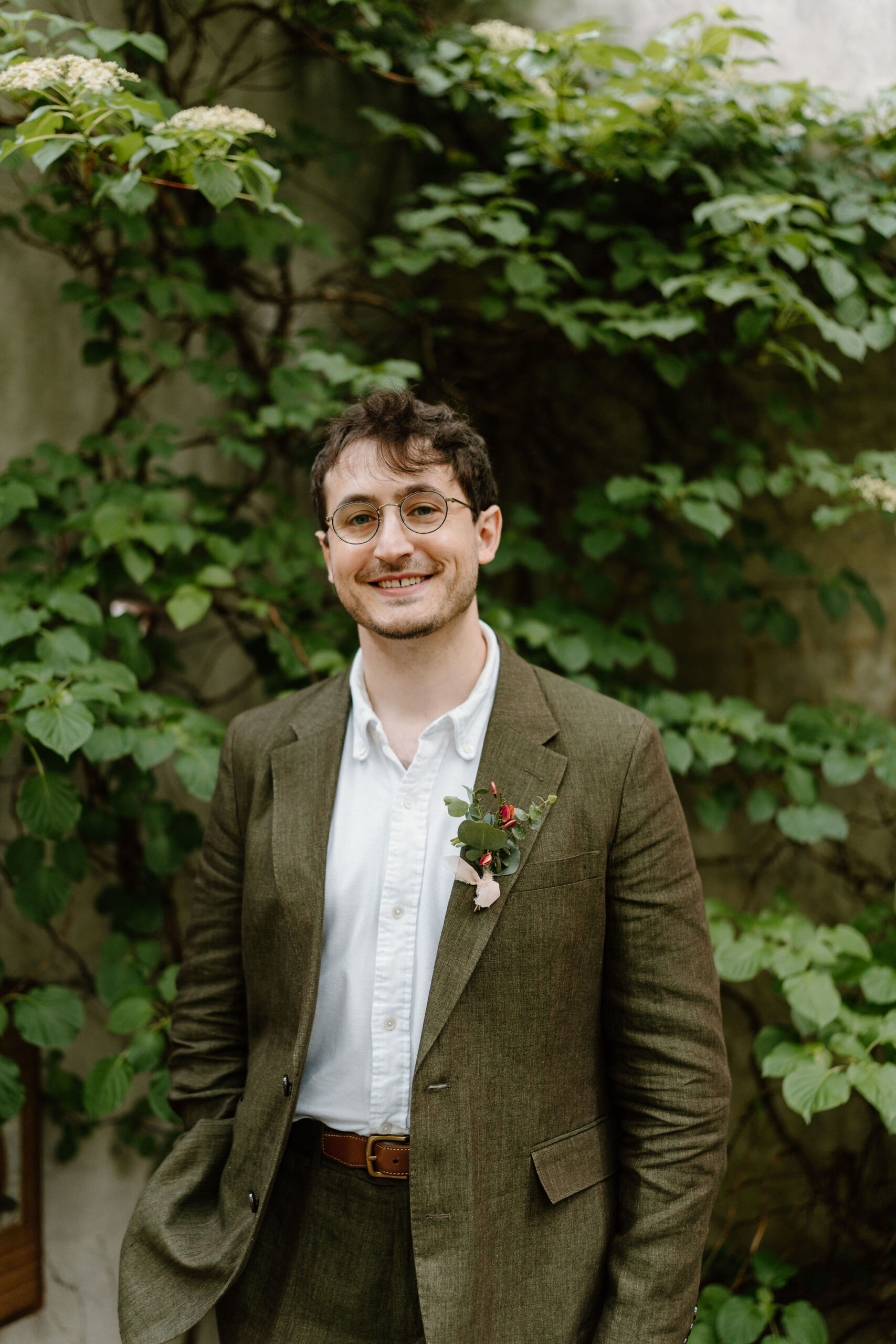 groom (brown hair, olive green linen suit) smiling at the camera with one hand in his pocket, while wearing a boutoniere in front of a lush green wall