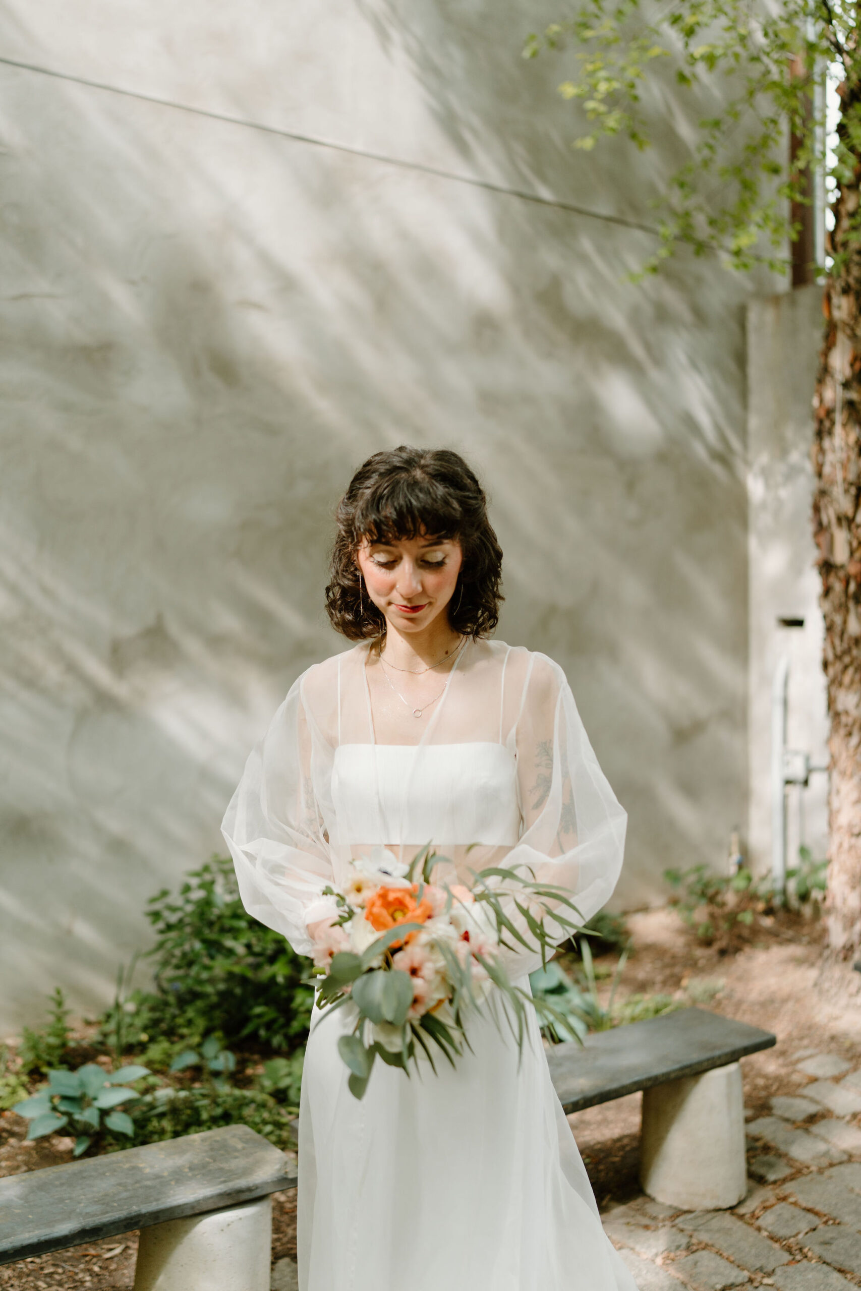 bride looking softly down at her bouquet held in front of her in front of a courtyard wall dappled with sunlight