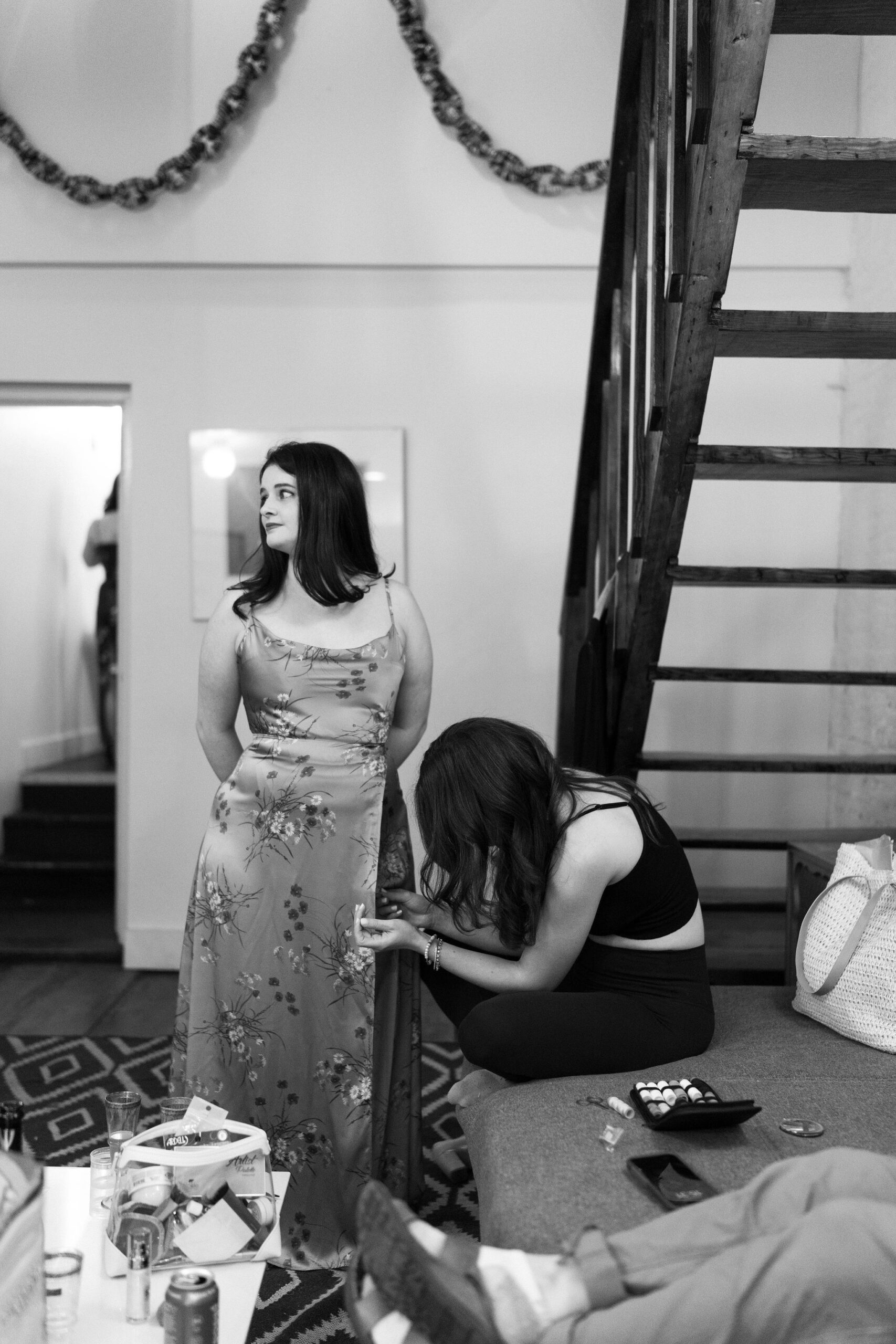 a woman in a floral dress, standing and having her dress sewn by another woman, seated on a couch, head down) while getting ready for a wedding in a cluttered room