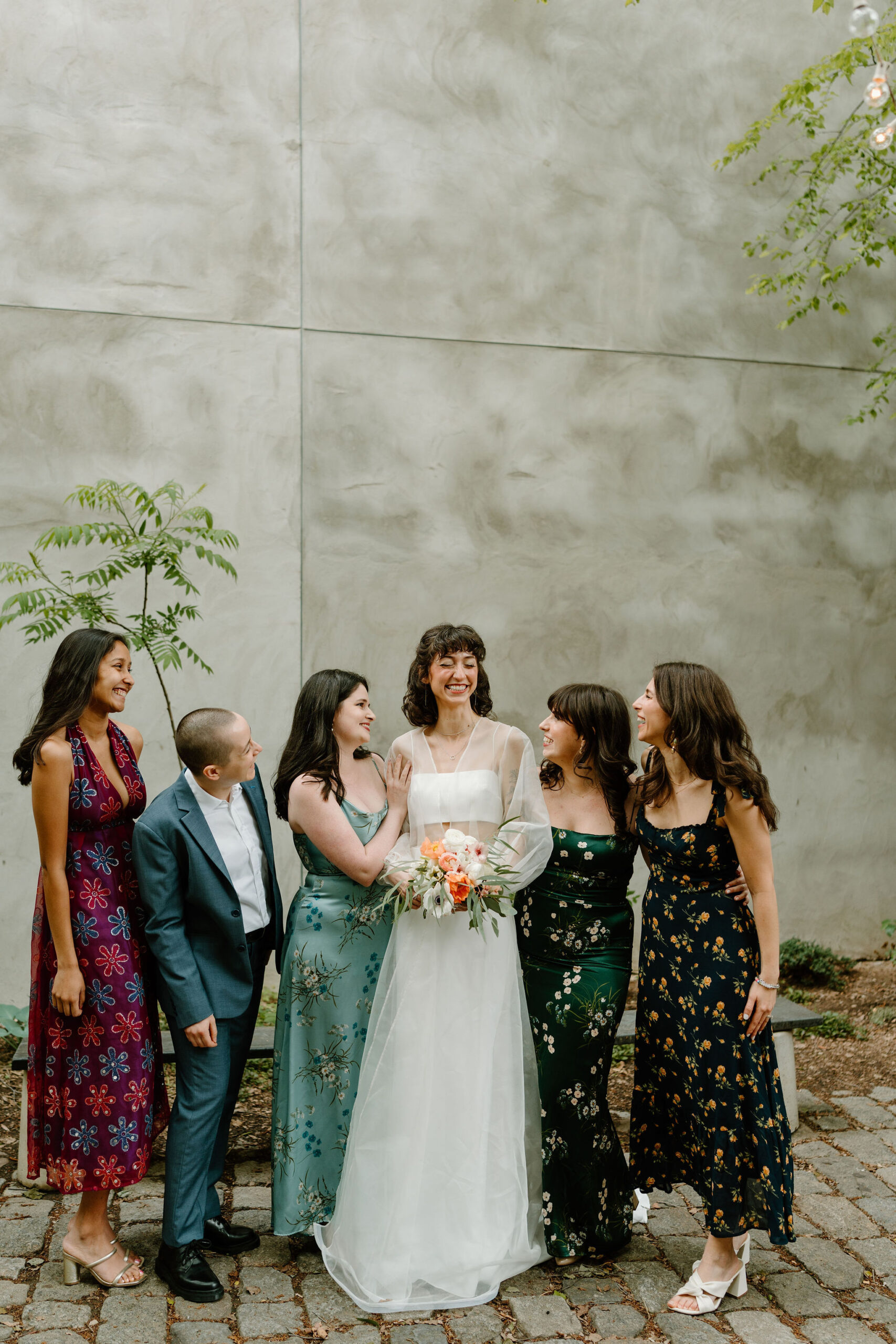 bride (wearing white) and five of her friends (in various colorful dresses and a blue suit) smiling at each other in a lush green cobblestone courtyard