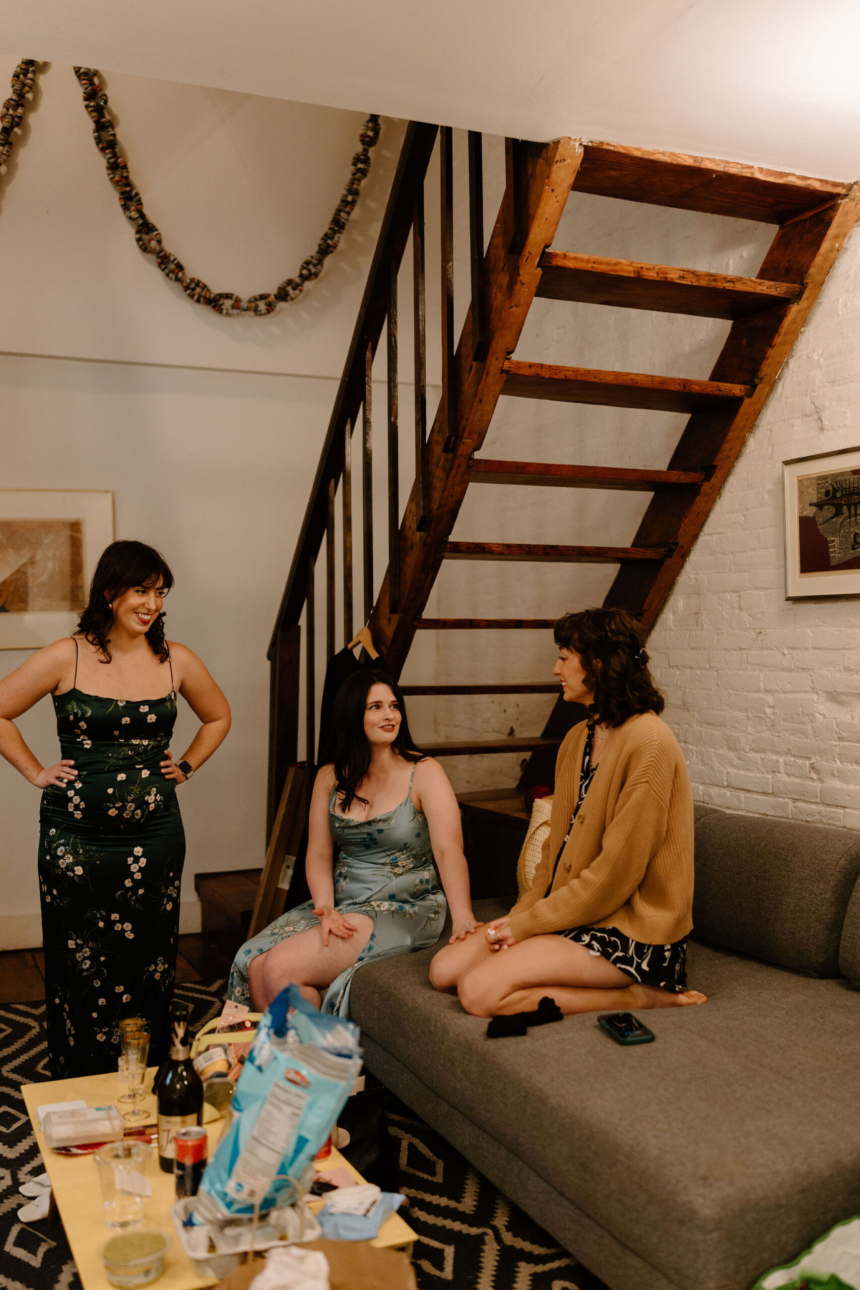 three friends in a cluttered room, one standing and two sitting on a couch, talking and getting ready for a wedding