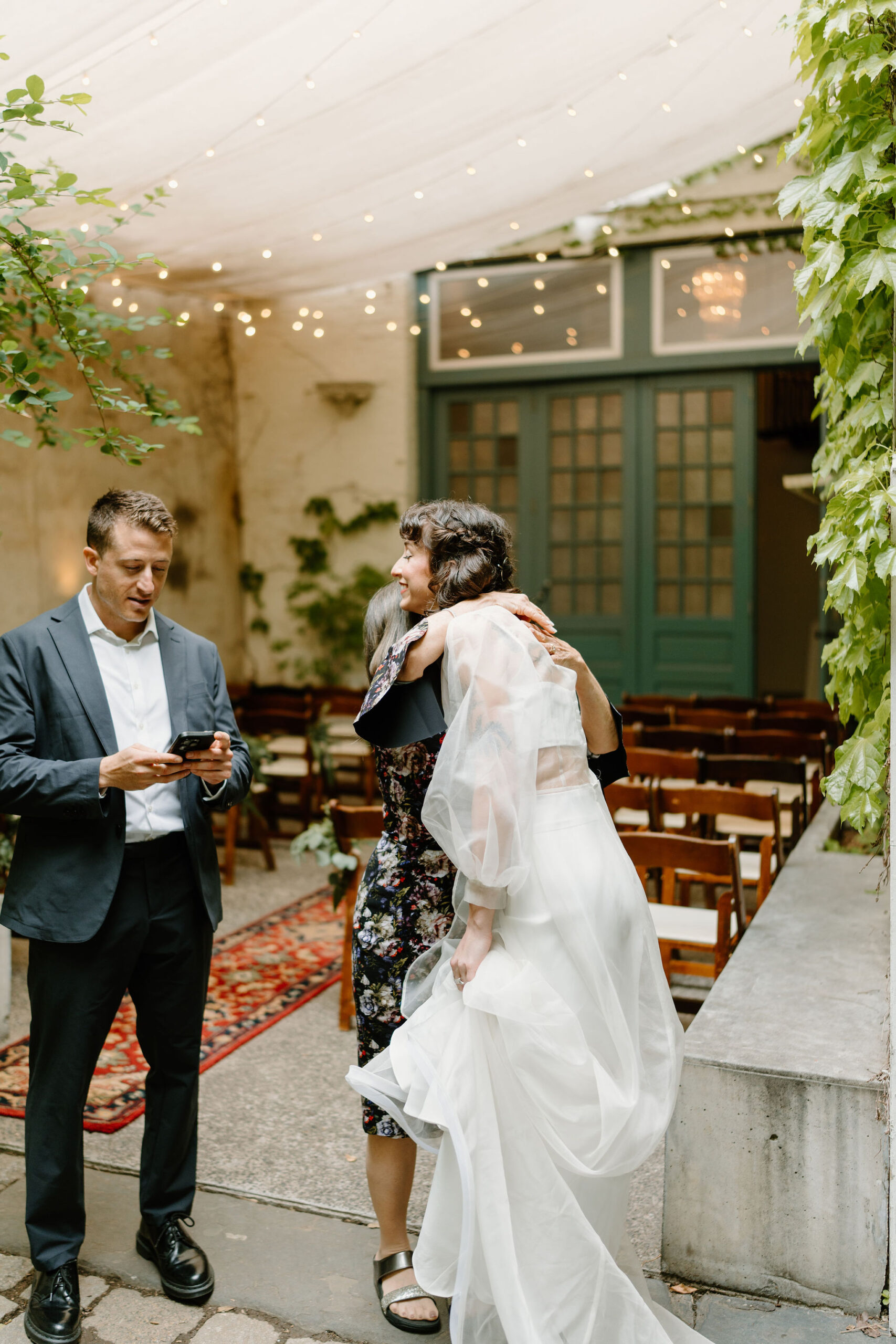 bride holding her skirt as she hugs her mother in law, their ceremony space in the background