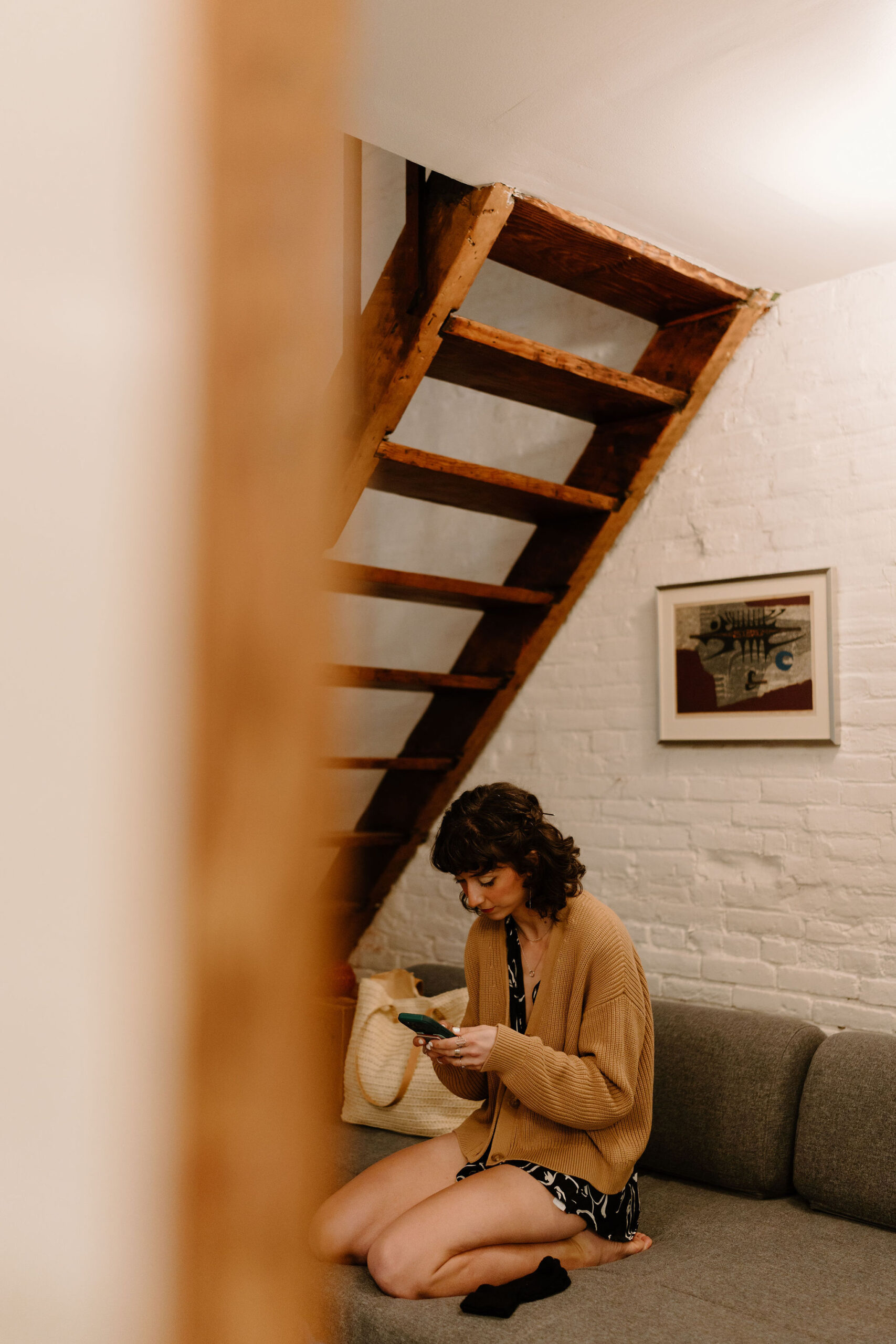 looking through a doorway, the bride is in her pajamas kneeling on the couch, looking at her phone