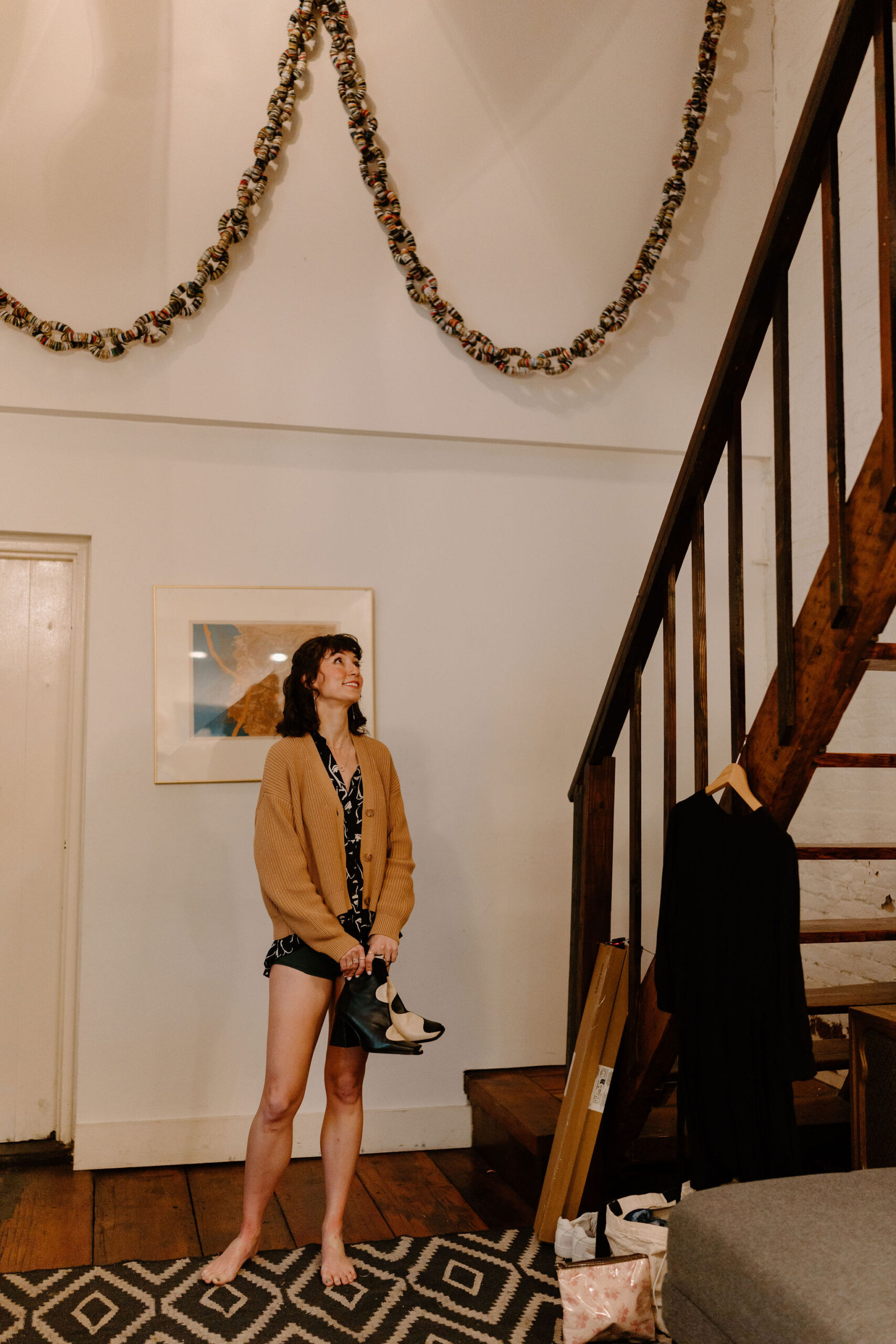 the bride, in her pajamas and holding her wedding boots, is looking up the stairs off-camera as she prepares to get dressed