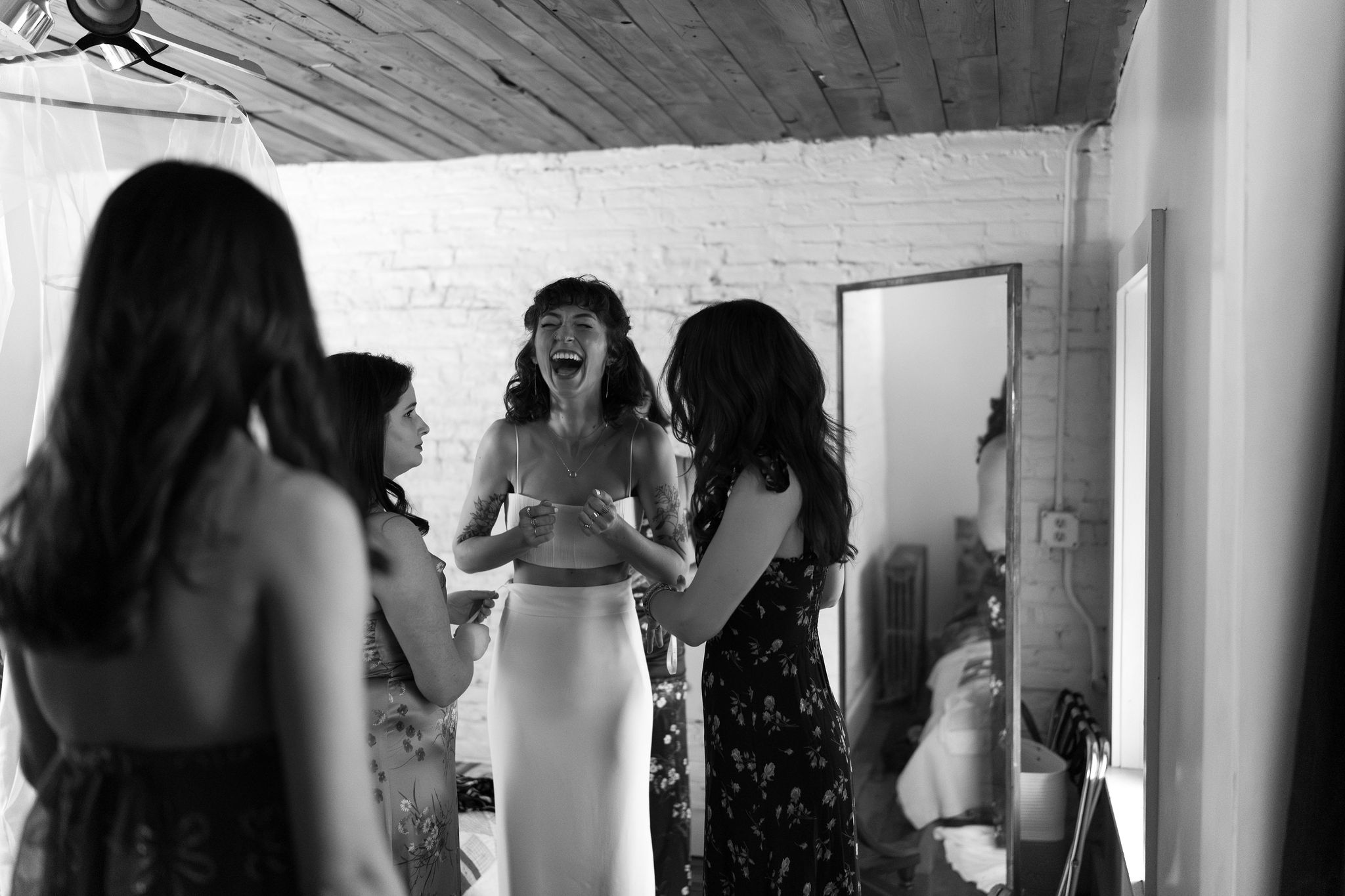 black and white image of a bride in a two piece wedding dress laughing, while four of her friends help her get into the dress