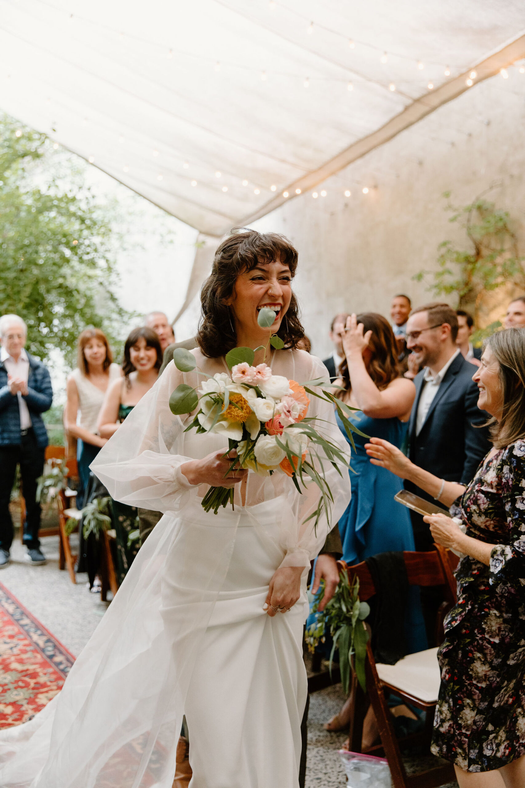 bride walking down the aisle to her ceremony, holding her flowers and laughing while her guests smile behind her