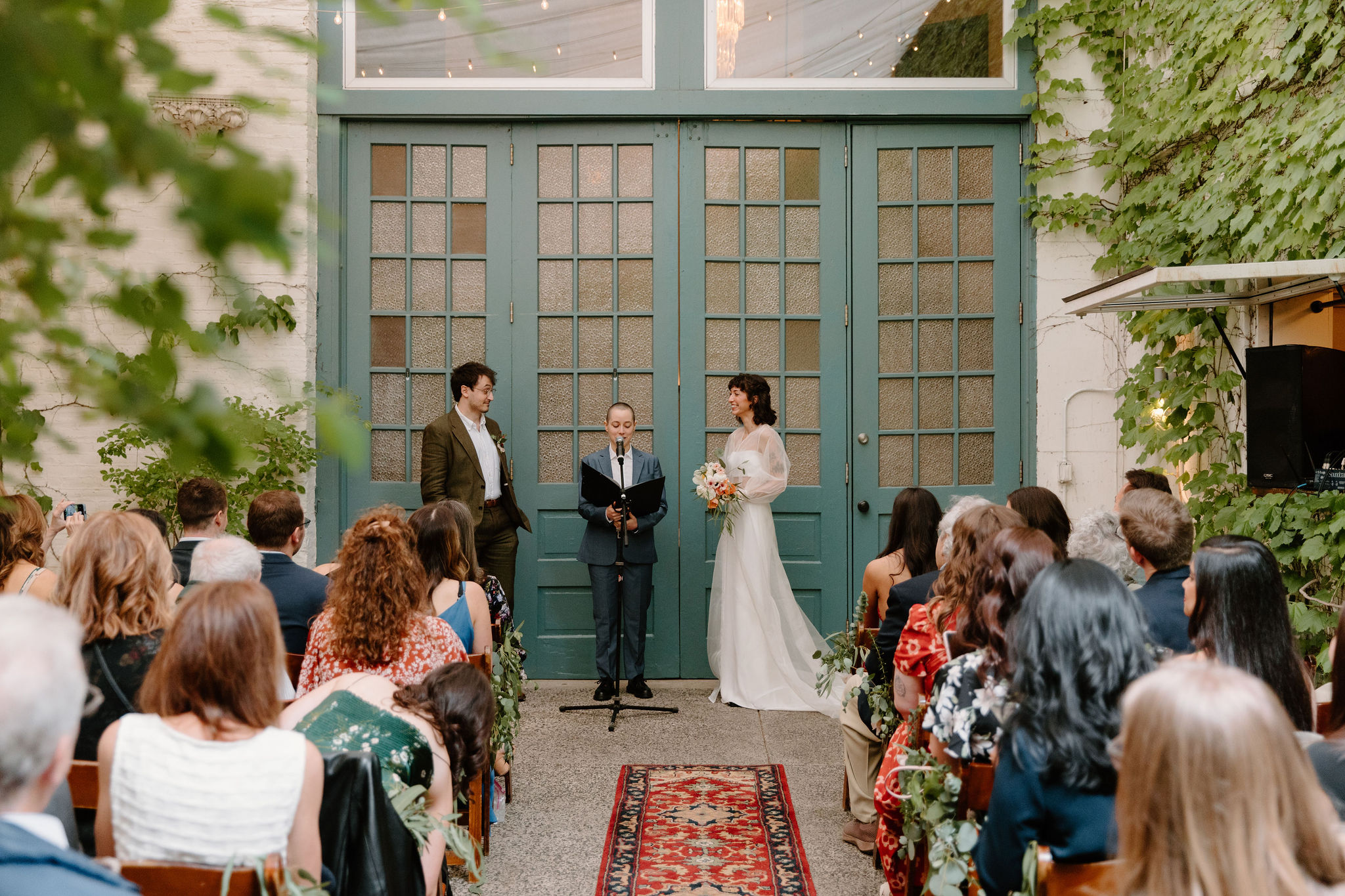 bride and groom being married by their friend, in front of large teal doors while their guests watch