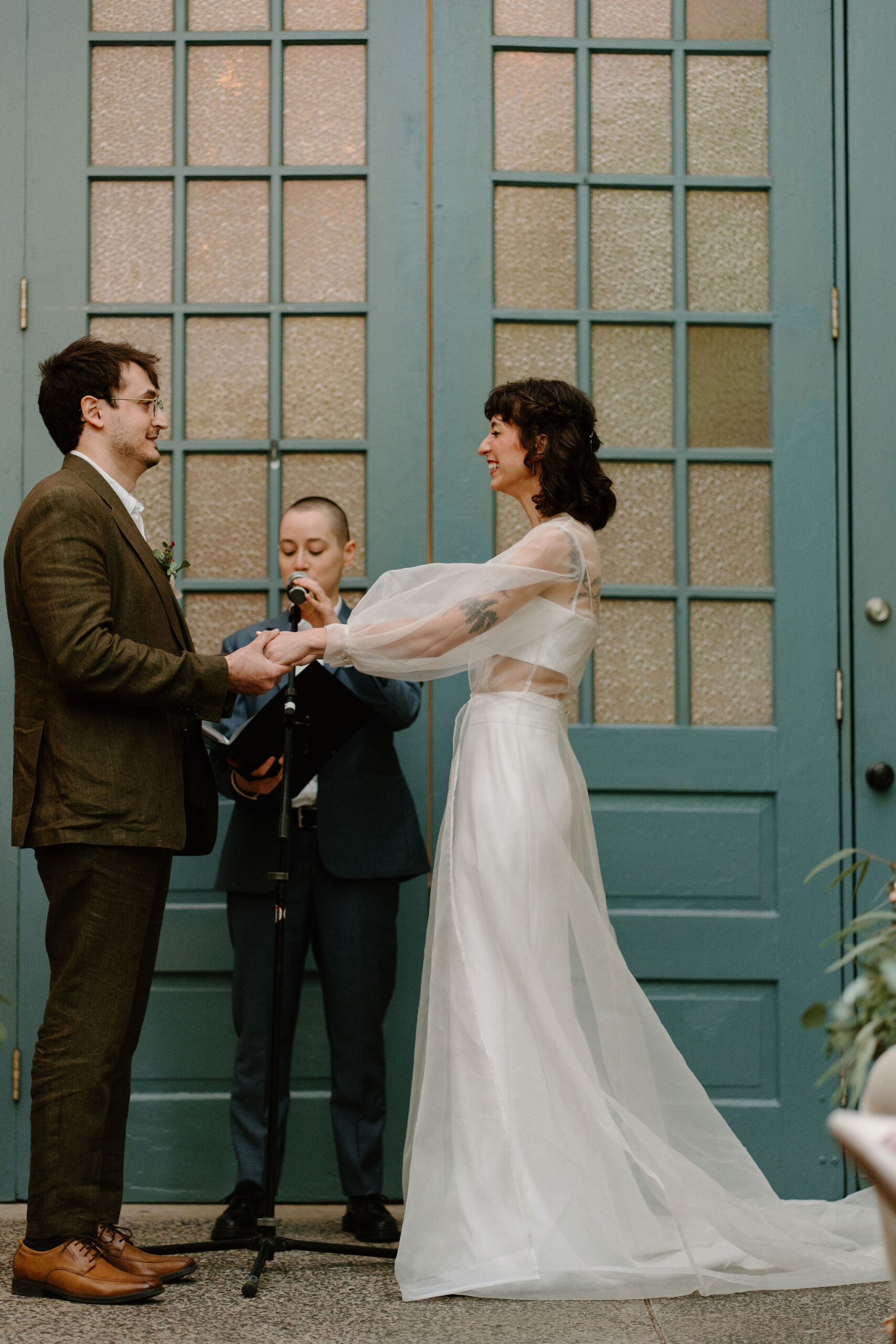 bride and groom being married by their friend, in front of large teal doors while their guests watch