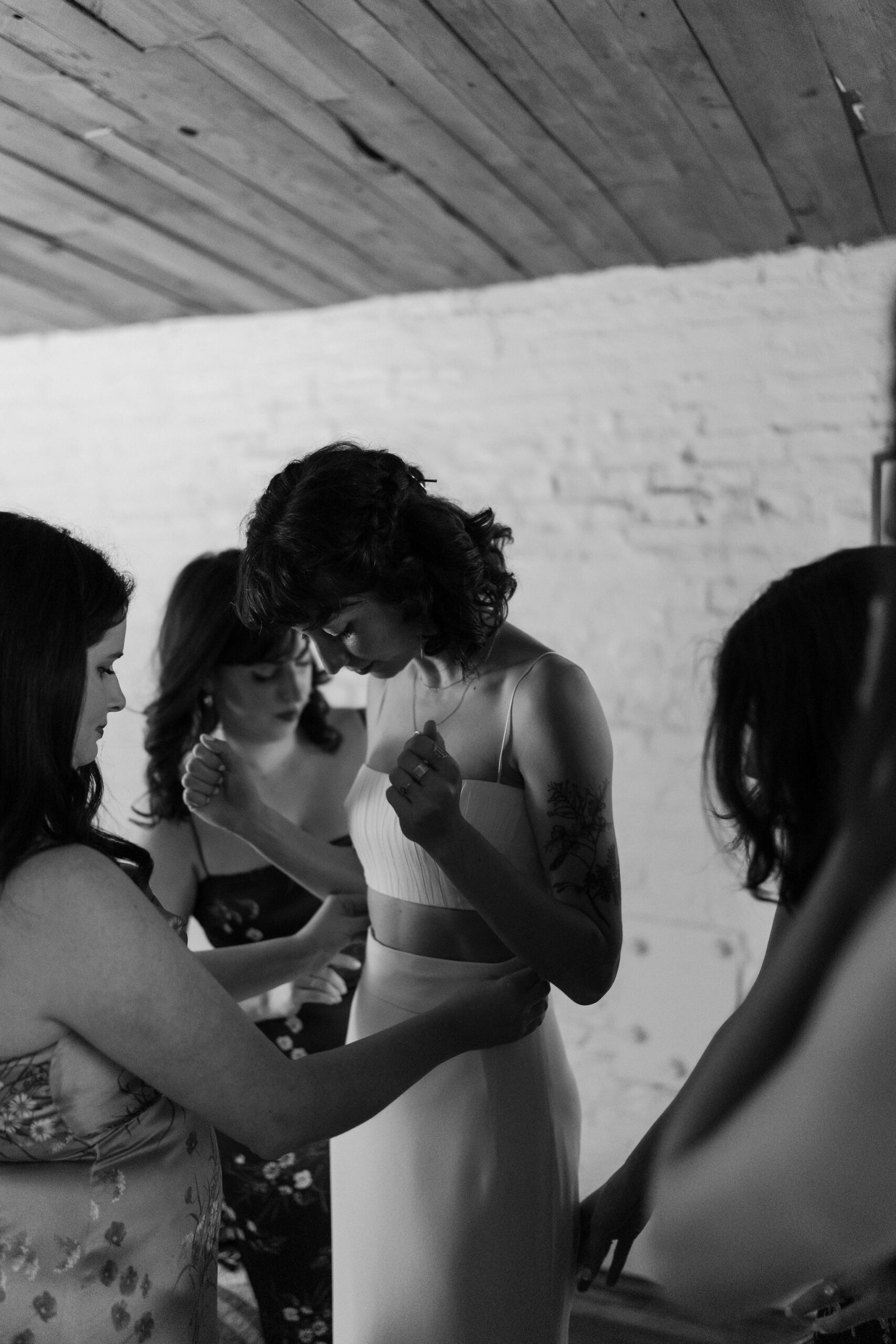 black and white image of a bride in a two piece dress with spaghetti straps, looking down at herself while her four friends fuss over the dress