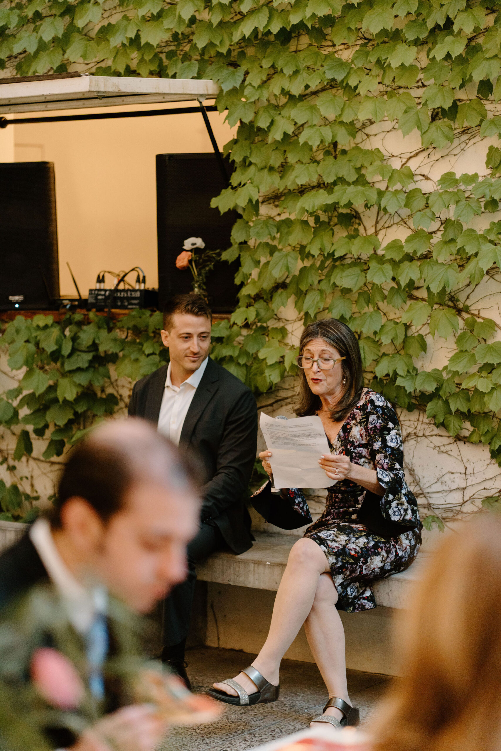 mother of the groom sitting on a bench, delivering a toast while guests sit and eat pizza