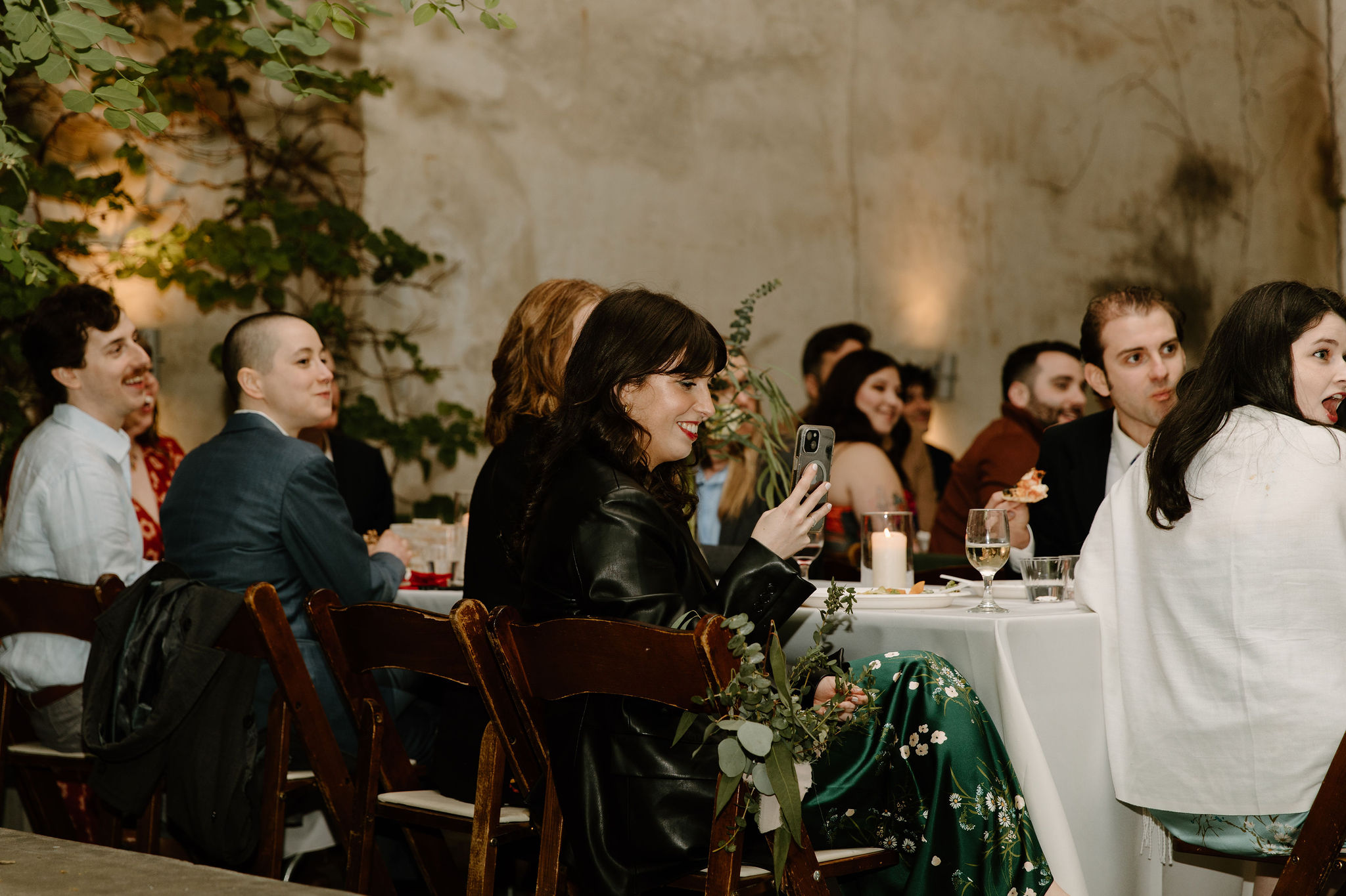 a guest smiling from her chair as she videos the speeches on her phone, other guests laughing around her