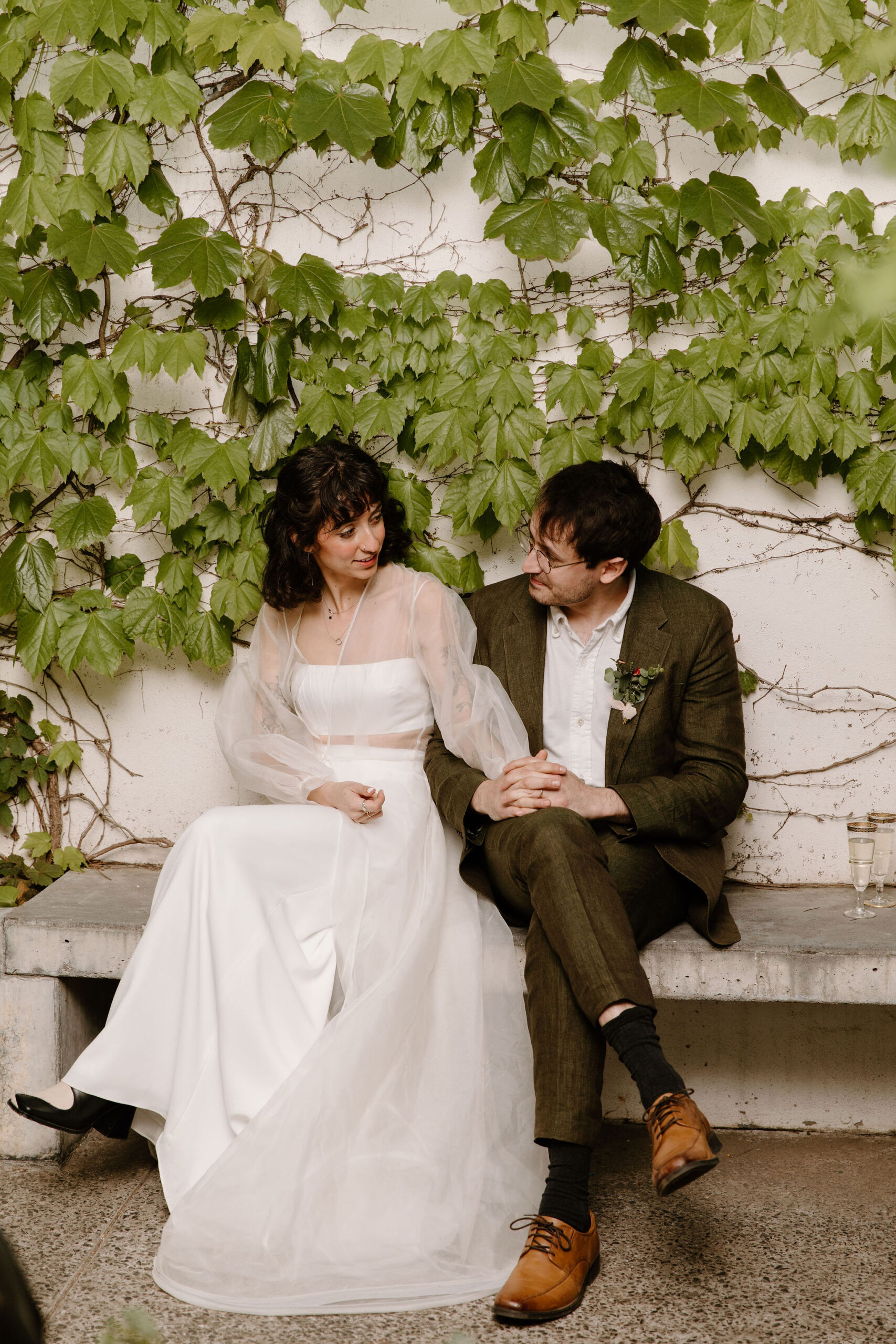 bride and groom sitting on a bench in front of an ivy wall, holding hands and looking at each other during speeches