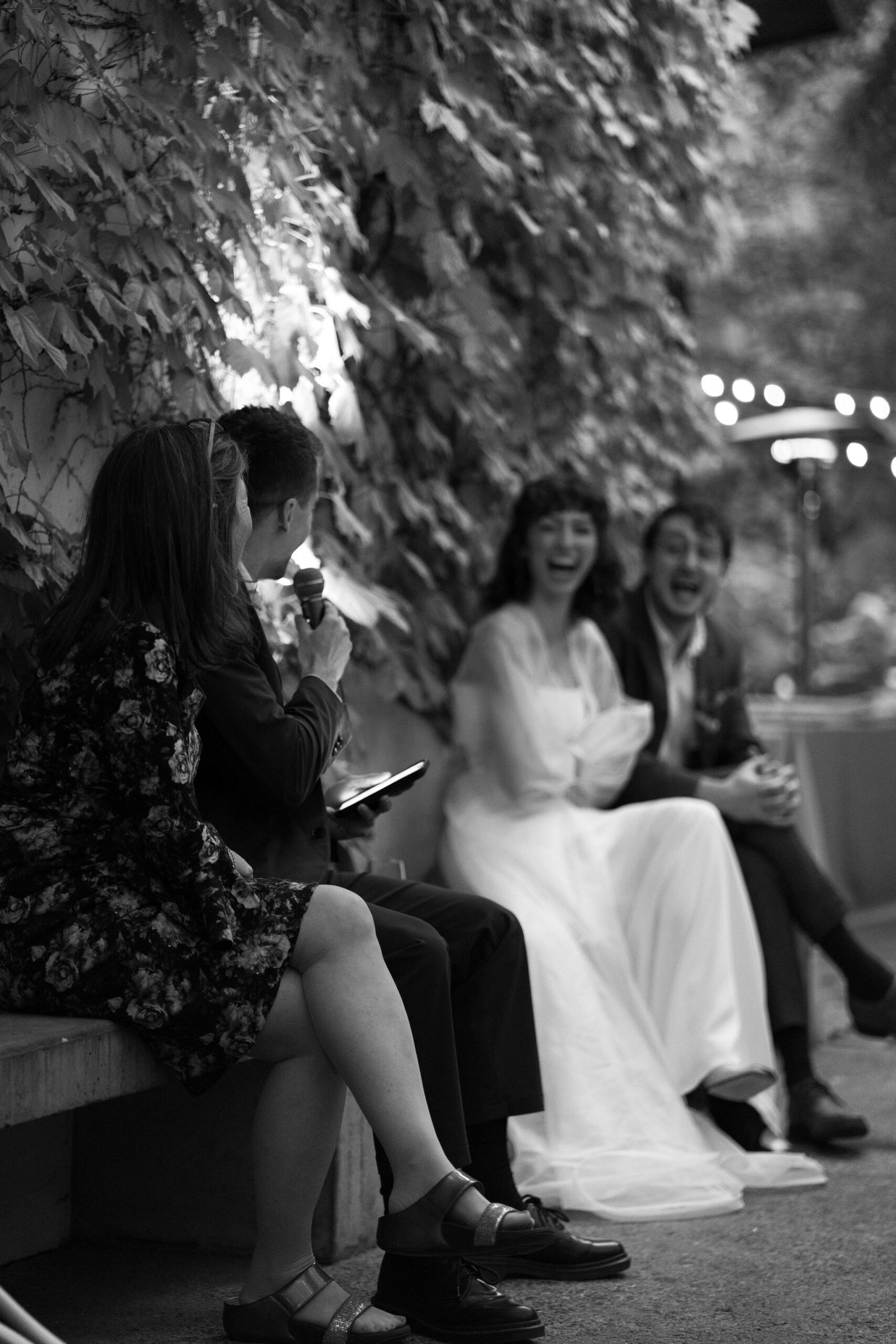 black and white image of brother of the groom, seated and delivering his speech to the bride and groom, laughing in the background