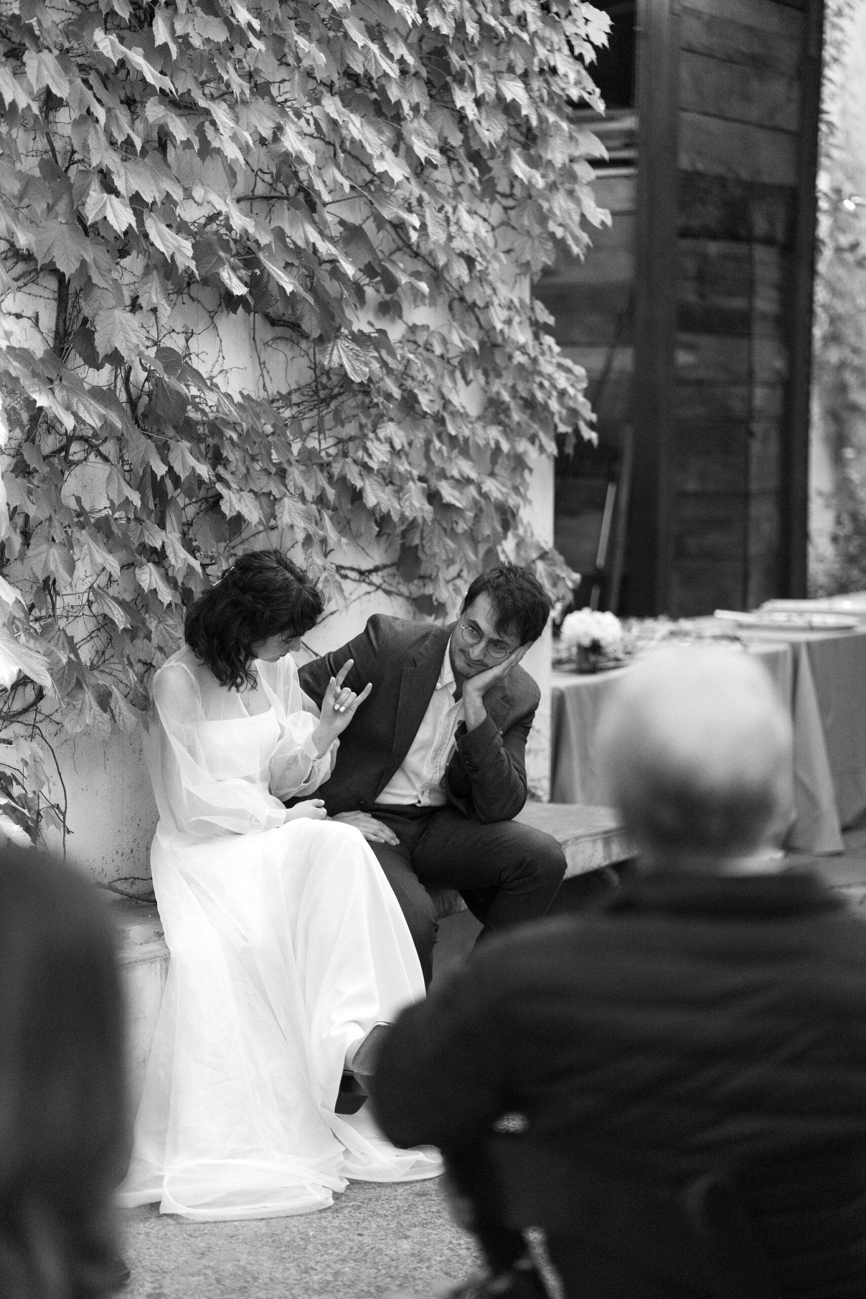 black and white image of bride and groom listening to speeches, while the bride throws a "rock on" symbol with her hands