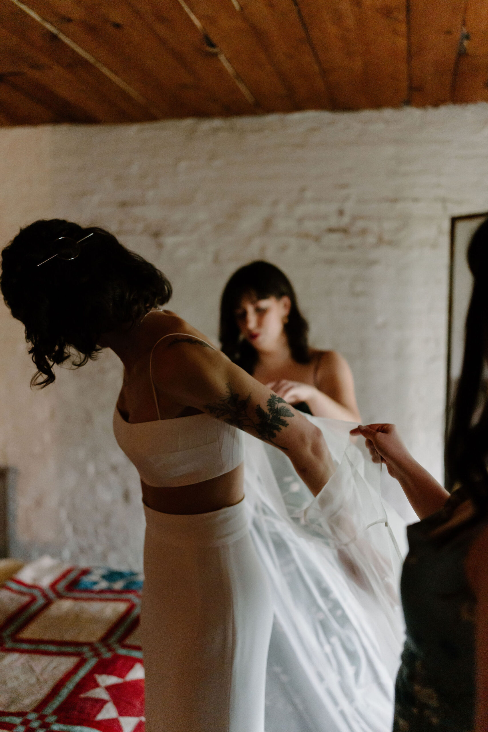 bride in a two piece wedding dress, putting on a sheer cape with help from two of her friends