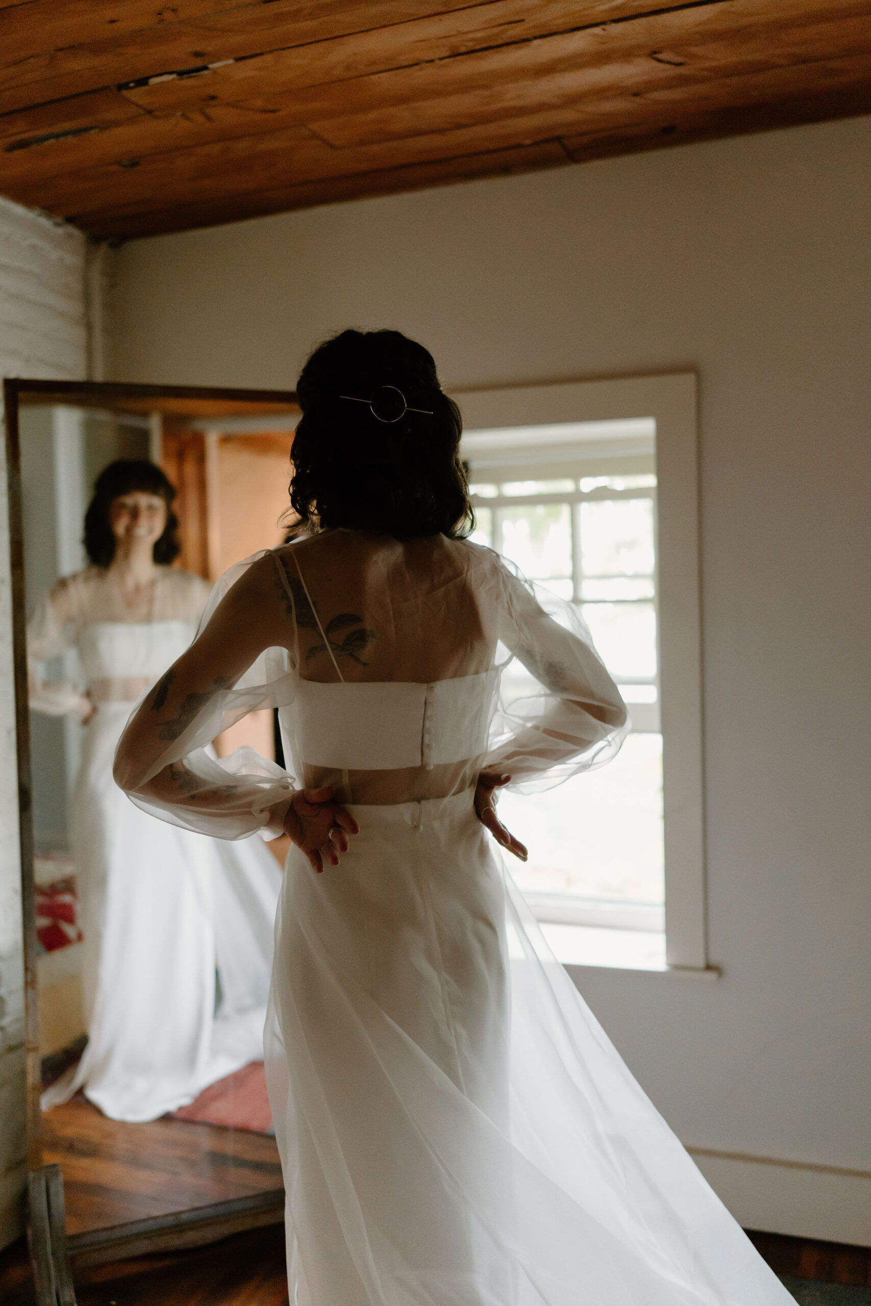 bride wearing a two piece wedding dress and a sheer cape, looking at herself in the mirror with her hands on her hips