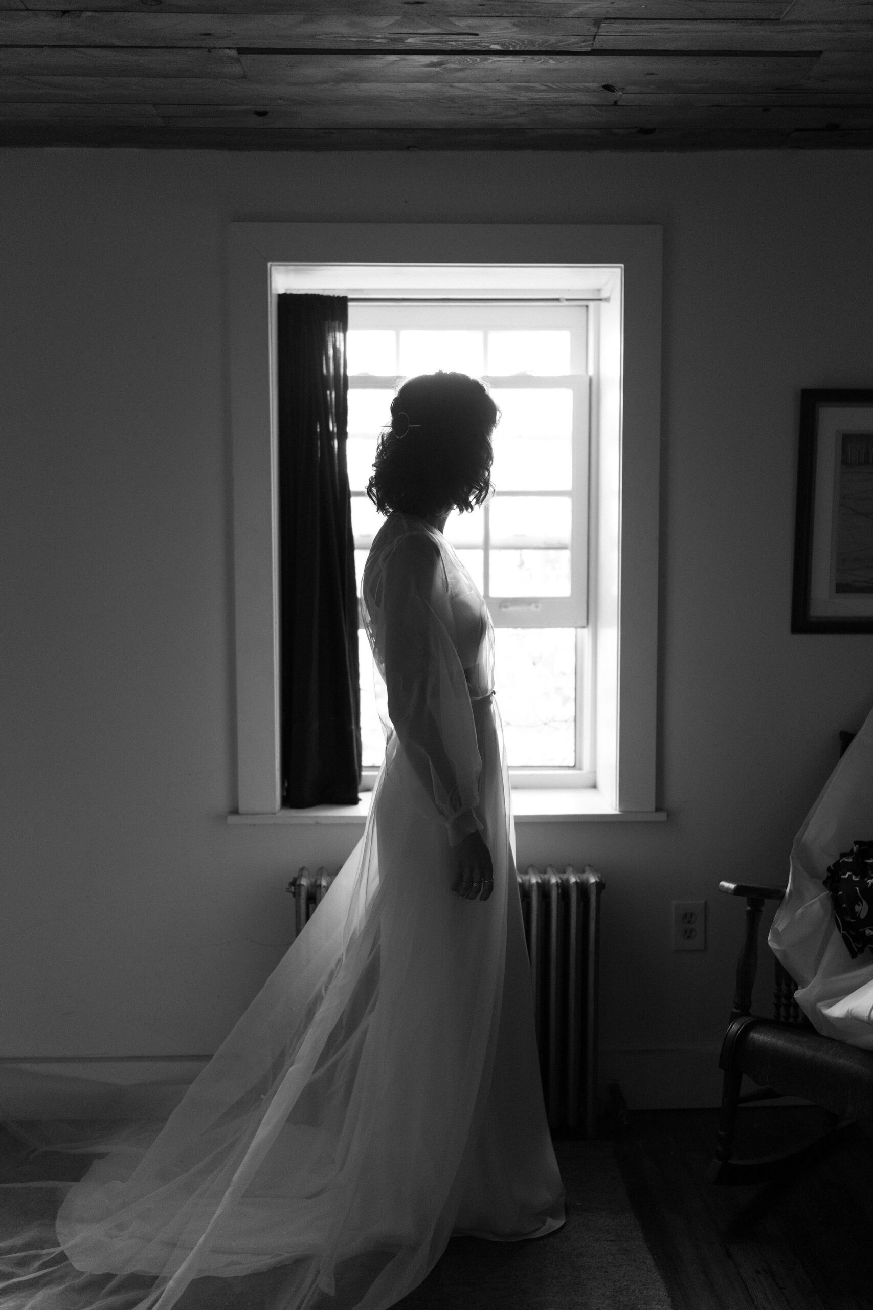 black and white image of a bride wearing a two piece wedding gown and a sheer cape, silhouetted looking out a window away from the camera