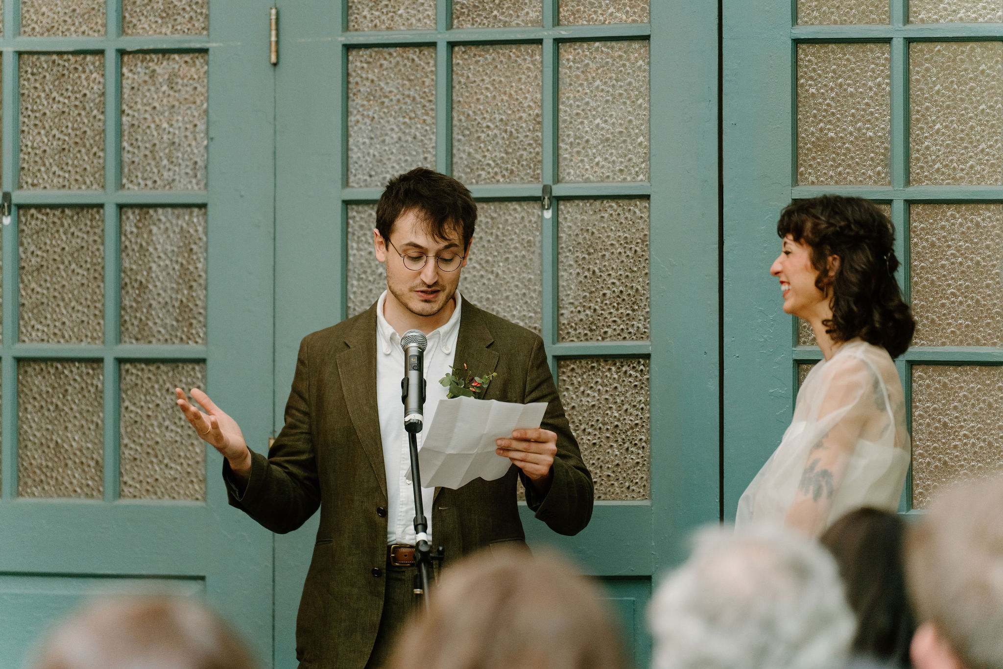 bride and groom exchanging vows during their ceremony, in front of large teal doors while their guests watch