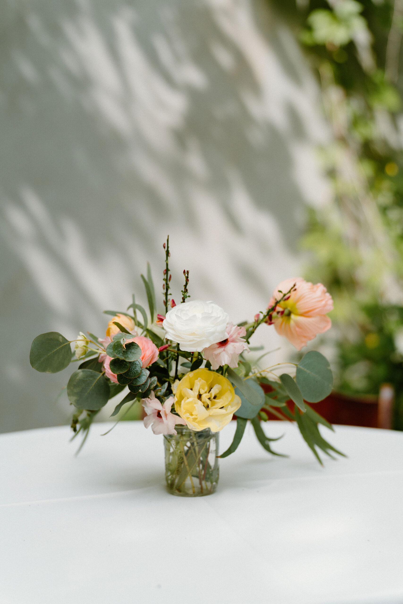 a small centerpiece flower vase holding poppies, peonies, and eucalyptus on a white tablecloth, outdoors against a white wall with lots of midday shadows across it