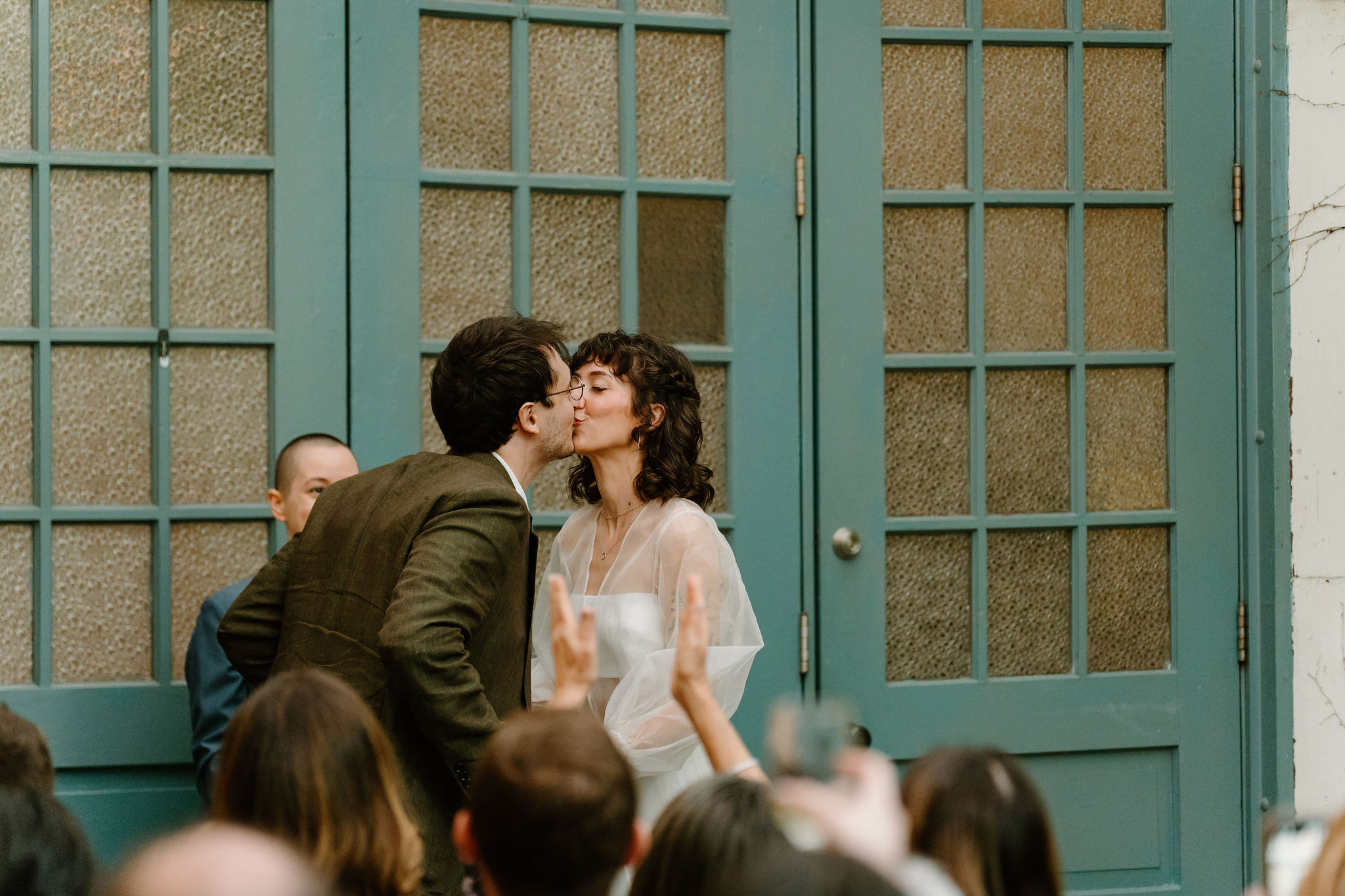 bride and groom exchanging their first kiss, in front of large teal doors while their guests watch